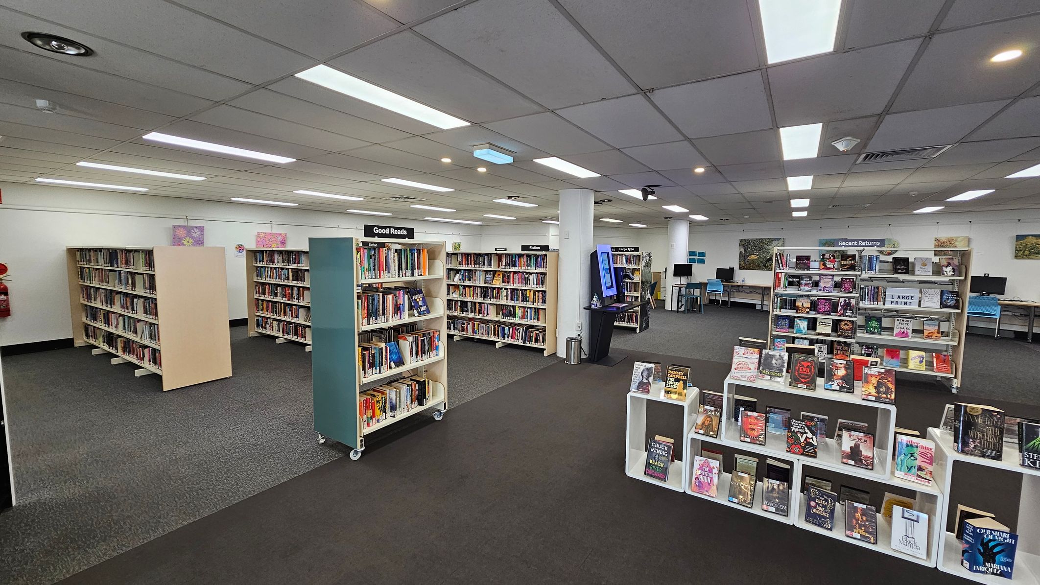 A library interior with several bookshelves filled with books. On the left, there are shelves labelled "Good Reads" and "Fiction". In the centre, a display features books on white stands. To the right, a "Recent Returns" section is visible. The background shows computer stations along the wall. The floor is carpeted in grey, and the ceiling has fluorescent lighting. A library interior with several bookshelves filled with books. On the left, there are shelves labelled "Good Reads" and "Fiction". In the centre, a display features books on white stands. To the right, a "Recent Returns" section is visible. The background shows computer stations along the wall. The floor is carpeted in grey, and the ceiling has fluorescent lighting.