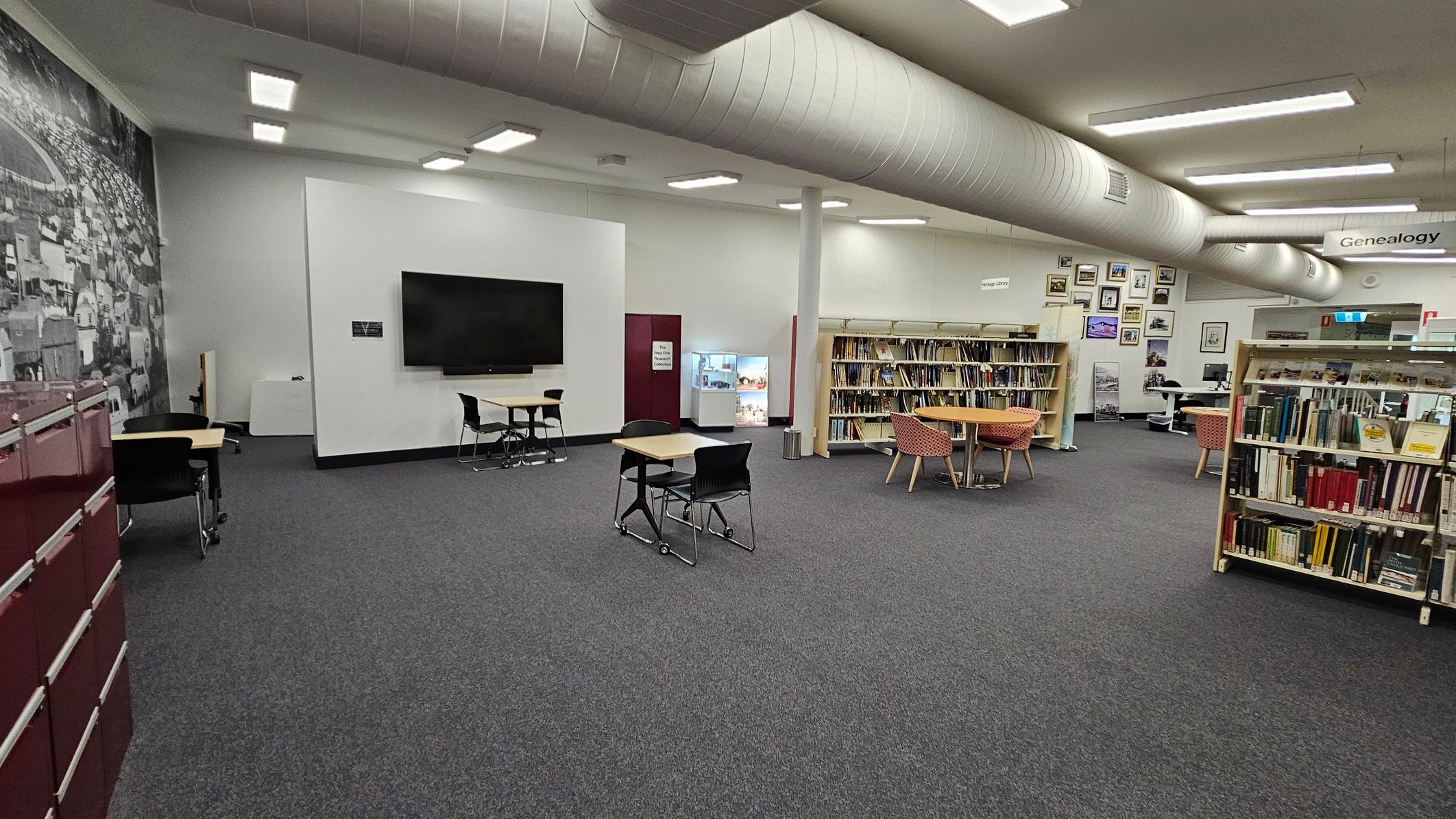 A room with a red wall featuring the word "Heritage" and arrows pointing towards an open doorway. To the left, there's a bulletin board with various posters and a printer below it. In the background, several computer workstations with blue chairs are lined up against a white wall. The floor is carpeted, and the ceiling has fluorescent lights. A small table with two blue chairs is on the right. A room with a red wall featuring the word "Heritage" and arrows pointing towards an open doorway. To the left, there's a bulletin board with various posters and a printer below it. In the background, several computer workstations with blue chairs are lined up against a white wall. The floor is carpeted, and the ceiling has fluorescent lights. A small table with two blue chairs is on the right.