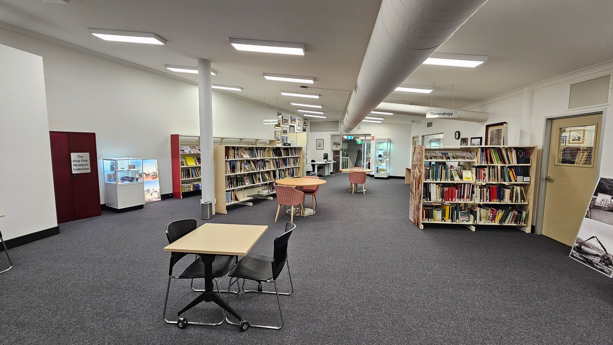 A library interior with grey carpeted flooring. On the left, there is a display case and a section labelled "The Fred Fink Research Collection." In the centre, there are bookshelves filled with books and a few round tables with red-patterned chairs. On the right, more bookshelves are visible, along with a door. The ceiling has fluorescent lights and a large ventilation duct running across. In the background, there are more tables and a computer station. A library interior with grey carpeted flooring. On the left, there is a display case and a section labelled "The Fred Fink Research Collection." In the centre, there are bookshelves filled with books and a few round tables with red-patterned chairs. On the right, more bookshelves are visible, along with a door. The ceiling has fluorescent lights and a large ventilation duct running across. In the background, there are more tables and a computer station.