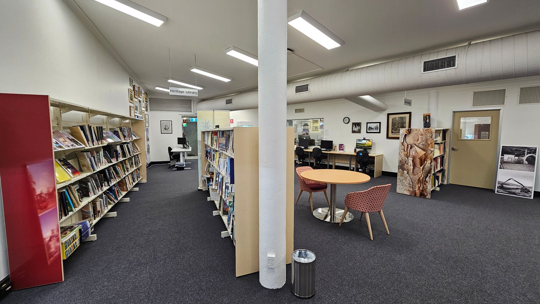 A library interior with a central white pillar. On the left, there are bookshelves filled with books and a red panel. In the middle, more bookshelves are visible. On the right, there's a round table with two chairs, a computer workstation with chairs, and framed pictures on the wall. A door with a window is also on the right. The floor is carpeted, and the ceiling has fluorescent lights and a large air duct. A small bin is near the pillar. A library interior with a central white pillar. On the left, there are bookshelves filled with books and a red panel. In the middle, more bookshelves are visible. On the right, there's a round table with two chairs, a computer workstation with chairs, and framed pictures on the wall. A door with a window is also on the right. The floor is carpeted, and the ceiling has fluorescent lights and a large air duct. A small bin is near the pillar.