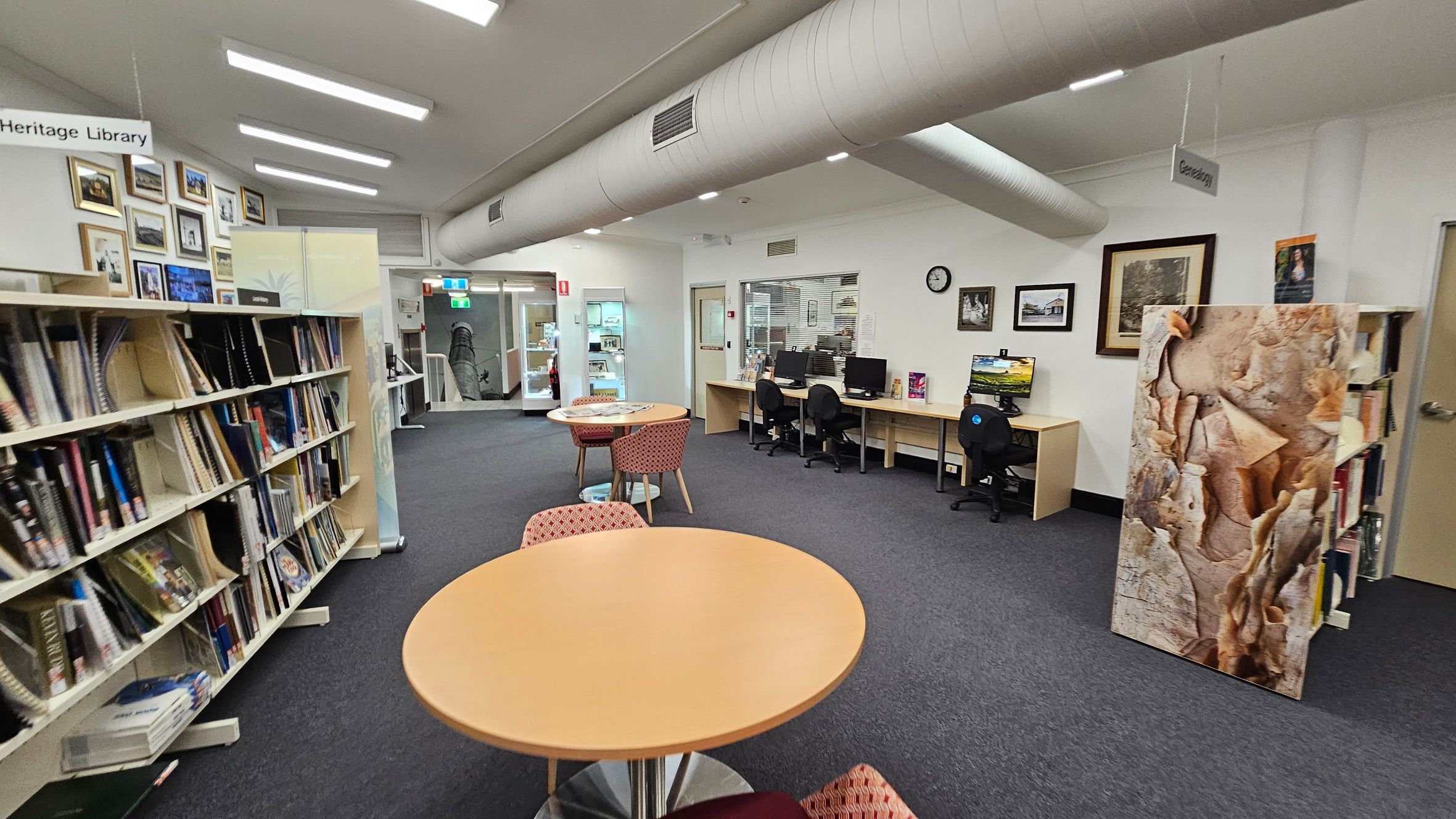 A library interior with bookshelves on the left filled with books and framed pictures above. In the centre, there are round tables with red-patterned chairs. On the right, a row of computer desks with chairs and a clock on the wall. A large artwork stands near the bookshelves. The floor is carpeted, and the ceiling has exposed ductwork and fluorescent lights. A sign reads "Heritage Library" on the left and "Genealogy" on the right. A library interior with bookshelves on the left filled with books and framed pictures above. In the centre, there are round tables with red-patterned chairs. On the right, a row of computer desks with chairs and a clock on the wall. A large artwork stands near the bookshelves. The floor is carpeted, and the ceiling has exposed ductwork and fluorescent lights. A sign reads "Heritage Library" on the left and "Genealogy" on the right.