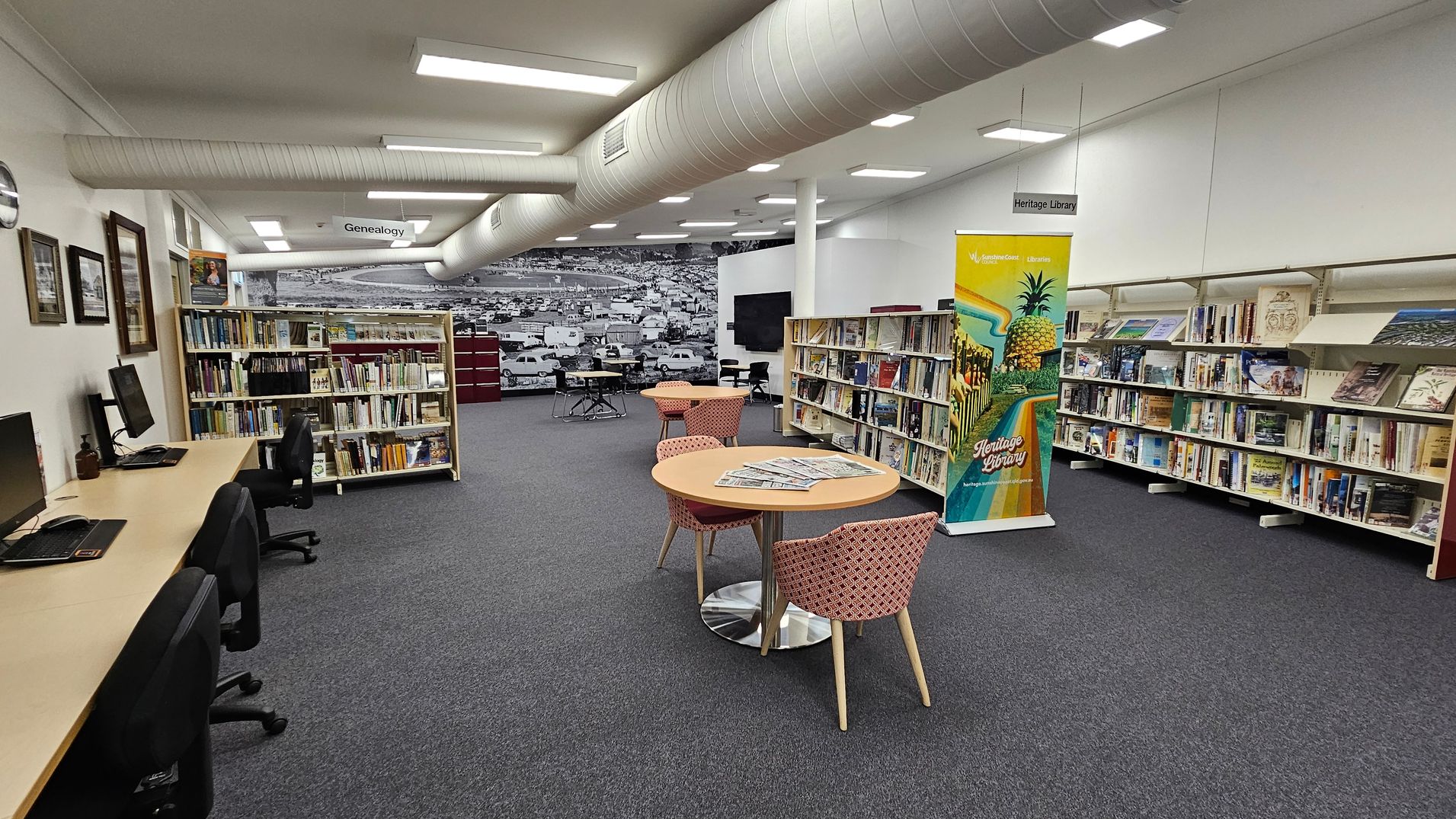 A library interior with a grey carpeted floor. On the left, there are computer desks with chairs and framed pictures on the wall. In the centre, round tables with patterned chairs hold newspapers. Bookshelves line the walls, filled with books. A colourful banner stands near the shelves, promoting the Heritage Library. The ceiling has exposed ductwork and fluorescent lighting. A large black-and-white mural of a townscape is visible in the background. A library interior with a grey carpeted floor. On the left, there are computer desks with chairs and framed pictures on the wall. In the centre, round tables with patterned chairs hold newspapers. Bookshelves line the walls, filled with books. A colourful banner stands near the shelves, promoting the Heritage Library. The ceiling has exposed ductwork and fluorescent lighting. A large black-and-white mural of a townscape is visible in the background.