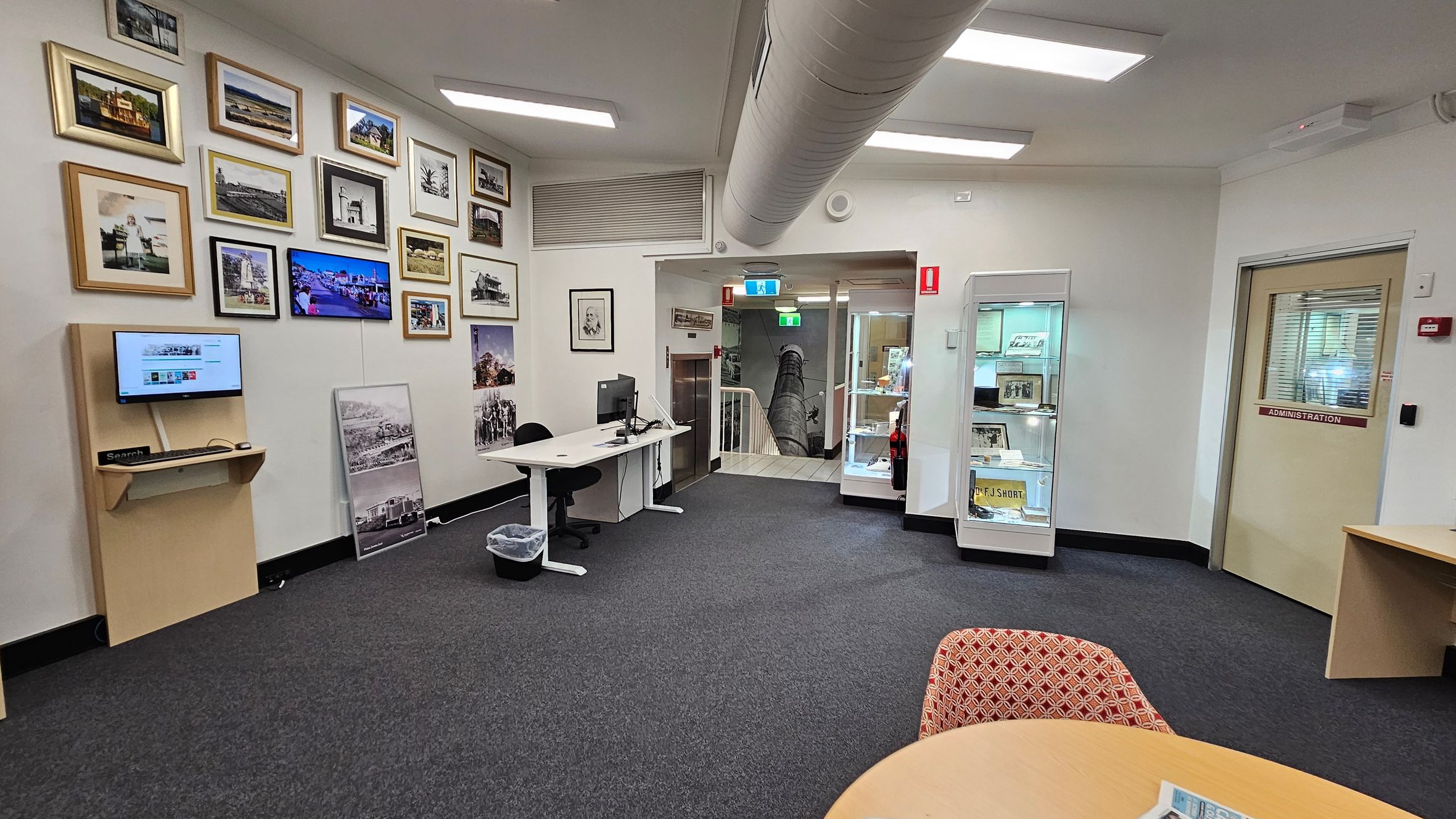 A room with a grey carpet and white walls. On the left, a wall displays numerous framed photographs and a small monitor on a wooden shelf. Below, a desk with a computer and chair. In the centre, a doorway leads to a staircase, with a glass display cabinet nearby. On the right, a door marked "Administration" and a round table with a patterned chair. The ceiling has fluorescent lights and a large air duct. A room with a grey carpet and white walls. On the left, a wall displays numerous framed photographs and a small monitor on a wooden shelf. Below, a desk with a computer and chair. In the centre, a doorway leads to a staircase, with a glass display cabinet nearby. On the right, a door marked "Administration" and a round table with a patterned chair. The ceiling has fluorescent lights and a large air duct.