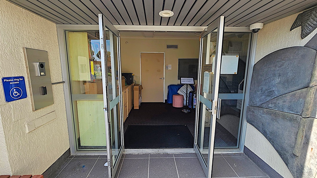 Glass double doors are open, leading into a building. On the left, there's a wall-mounted intercom system with a blue sign indicating wheelchair access. Inside, a beige door is visible at the back, with a small table and some items to the left. On the right, a decorative wall feature is partially visible. The ceiling is lined with grey panels, and a security camera is mounted near the entrance. The floor is tiled in dark grey. Glass double doors are open, leading into a building. On the left, there's a wall-mounted intercom system with a blue sign indicating wheelchair access. Inside, a beige door is visible at the back, with a small table and some items to the left. On the right, a decorative wall feature is partially visible. The ceiling is lined with grey panels, and a security camera is mounted near the entrance. The floor is tiled in dark grey.