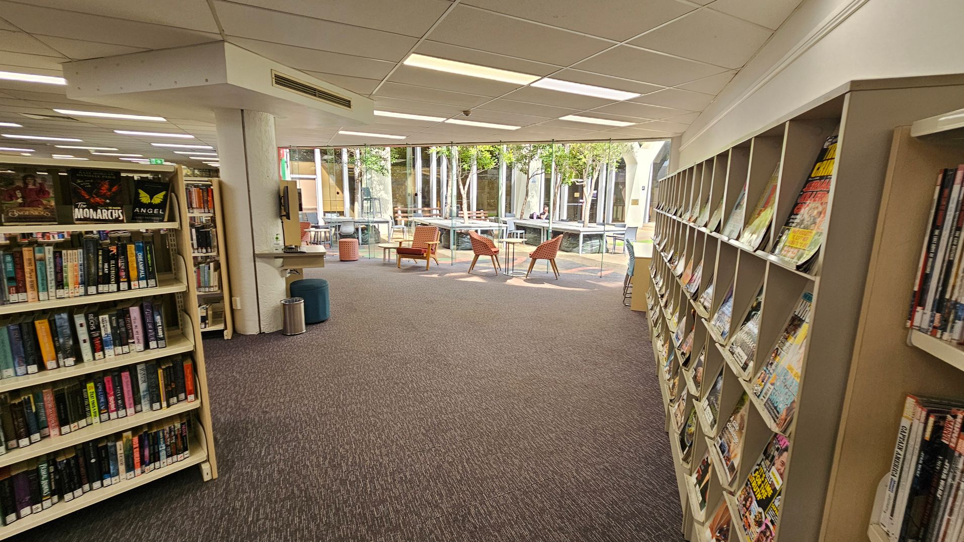 A library interior with bookshelves on both sides. On the left, shelves are filled with books, including a visible title "Goddess Crown". On the right, shelves hold DVDs and magazines. The floor is carpeted in a dark colour, and the ceiling has rectangular light panels. In the background, large windows reveal an outdoor area with trees and a few chairs and tables. A library interior with bookshelves on both sides. On the left, shelves are filled with books, including a visible title "Goddess Crown". On the right, shelves hold DVDs and magazines. The floor is carpeted in a dark colour, and the ceiling has rectangular light panels. In the background, large windows reveal an outdoor area with trees and a few chairs and tables.