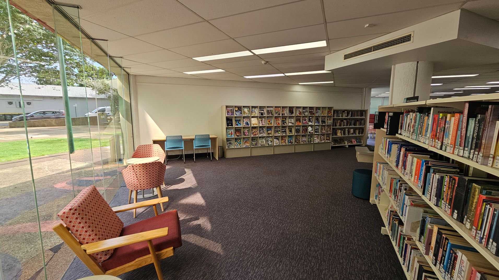 A library interior with a large glass window on the left, showing a view of the outside with grass and a road. Inside, there are two cushioned chairs and a small table near the window. Further back, two blue chairs are placed at a desk against the wall. The wall features shelves filled with magazines. On the right, there are bookshelves filled with books. The floor is carpeted in a dark colour, and the ceiling has rectangular lights. A library interior with a large glass window on the left, showing a view of the outside with grass and a road. Inside, there are two cushioned chairs and a small table near the window. Further back, two blue chairs are placed at a desk against the wall. The wall features shelves filled with magazines. On the right, there are bookshelves filled with books. The floor is carpeted in a dark colour, and the ceiling has rectangular lights.