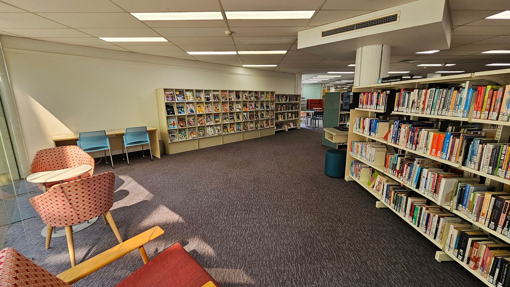 A library interior with bookshelves on the right filled with various books. In the middle, there are more shelves displaying magazines or comics. On the left, there are two blue chairs at a wooden desk and two red patterned chairs around a small round table. The floor is carpeted in a dark colour, and the ceiling has white panels with fluorescent lights. A library interior with bookshelves on the right filled with various books. In the middle, there are more shelves displaying magazines or comics. On the left, there are two blue chairs at a wooden desk and two red patterned chairs around a small round table. The floor is carpeted in a dark colour, and the ceiling has white panels with fluorescent lights.