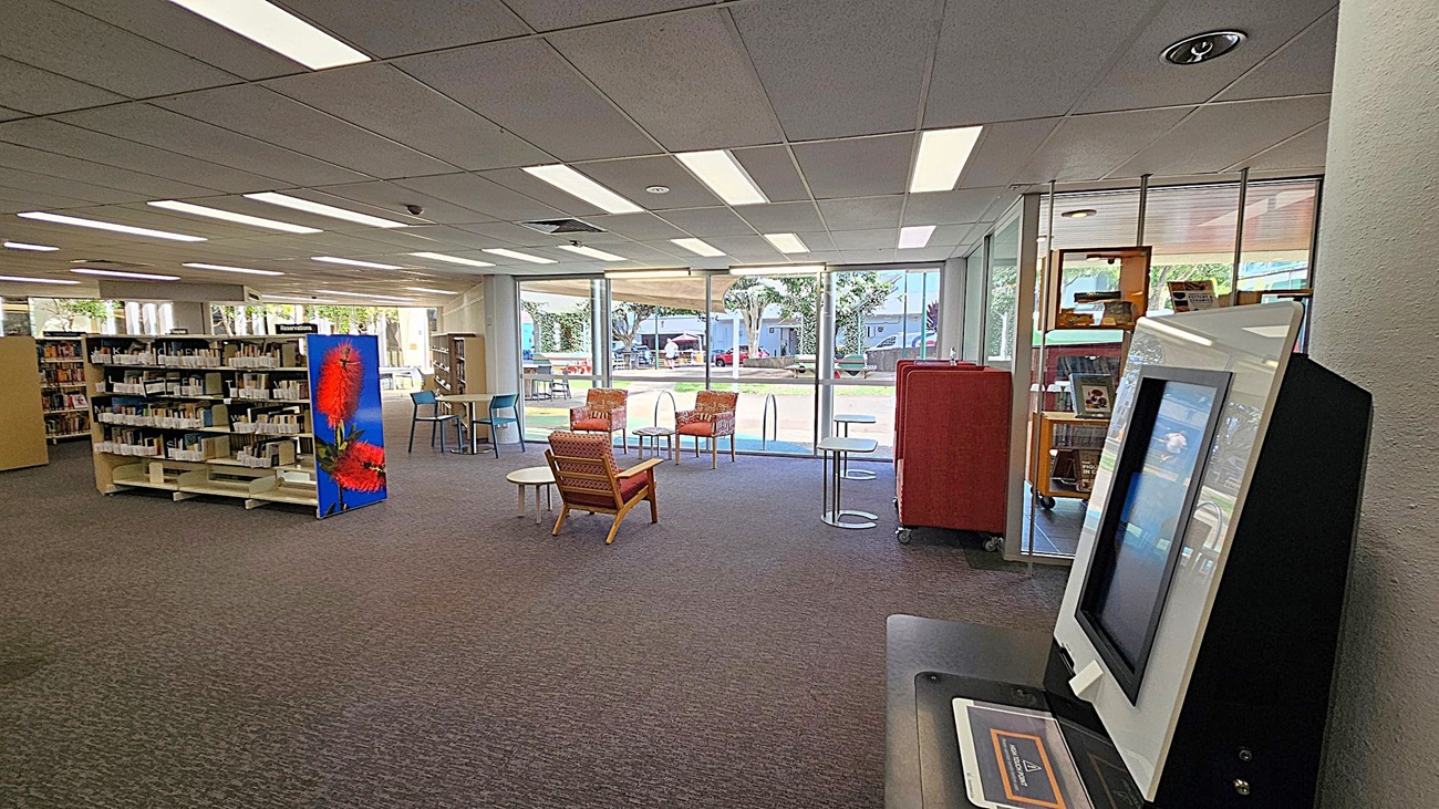 A library interior with a self-service kiosk on the right. In the centre, there are two orange chairs and a small round table on a carpeted floor. To the left, bookshelves are filled with books, and one end of a shelf features a large image of red flowers. Large windows in the background let in natural light, revealing an outdoor area with trees and a few people. The ceiling is lined with fluorescent lights. A library interior with a self-service kiosk on the right. In the centre, there are two orange chairs and a small round table on a carpeted floor. To the left, bookshelves are filled with books, and one end of a shelf features a large image of red flowers. Large windows in the background let in natural light, revealing an outdoor area with trees and a few people. The ceiling is lined with fluorescent lights.