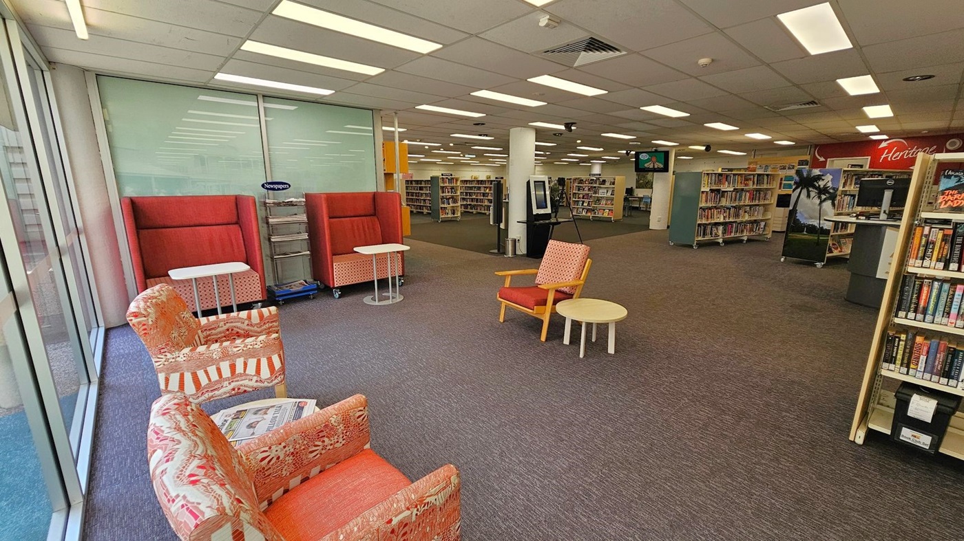 A library interior with a seating area on the left featuring patterned armchairs and small tables. Behind them are high-backed red chairs with additional tables. The floor is carpeted in a mix of grey and brown. In the centre, there are bookshelves filled with books, and a digital kiosk is visible. The ceiling has fluorescent lighting. On the right, more bookshelves and a computer station are present, with a sign reading "Heritage" in the background. A library interior with a seating area on the left featuring patterned armchairs and small tables. Behind them are high-backed red chairs with additional tables. The floor is carpeted in a mix of grey and brown. In the centre, there are bookshelves filled with books, and a digital kiosk is visible. The ceiling has fluorescent lighting. On the right, more bookshelves and a computer station are present, with a sign reading "Heritage" in the background.