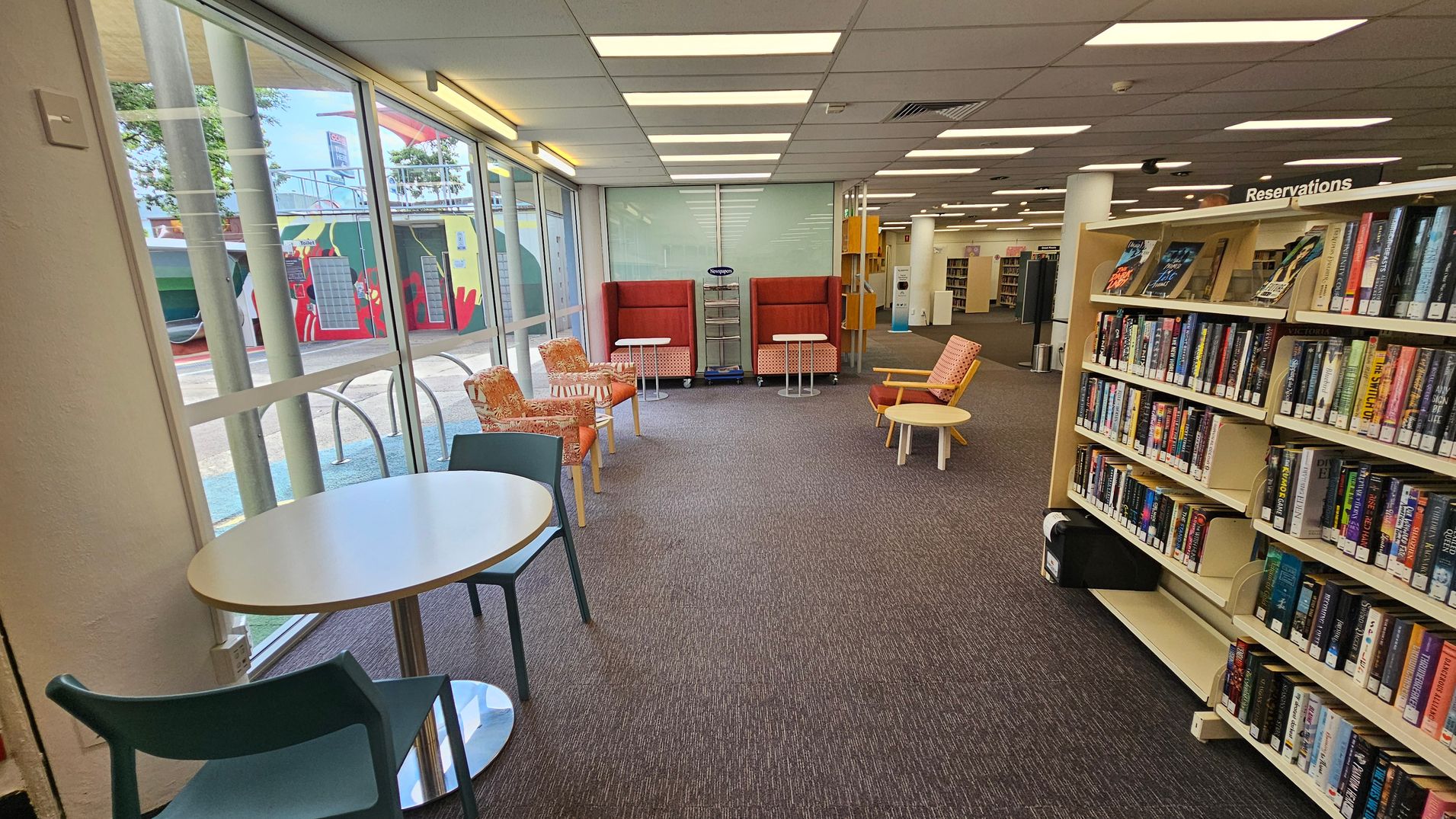 A library interior with a carpeted floor and a ceiling with fluorescent lights. On the left, large windows reveal an outdoor mural. In the centre, there are orange and patterned chairs with small round tables. On the right, a bookshelf labelled "Reservations" is filled with books. A small round table with a chair is near the window. The space is open and well-lit. A library interior with a carpeted floor and a ceiling with fluorescent lights. On the left, large windows reveal an outdoor mural. In the centre, there are orange and patterned chairs with small round tables. On the right, a bookshelf labelled "Reservations" is filled with books. A small round table with a chair is near the window. The space is open and well-lit.