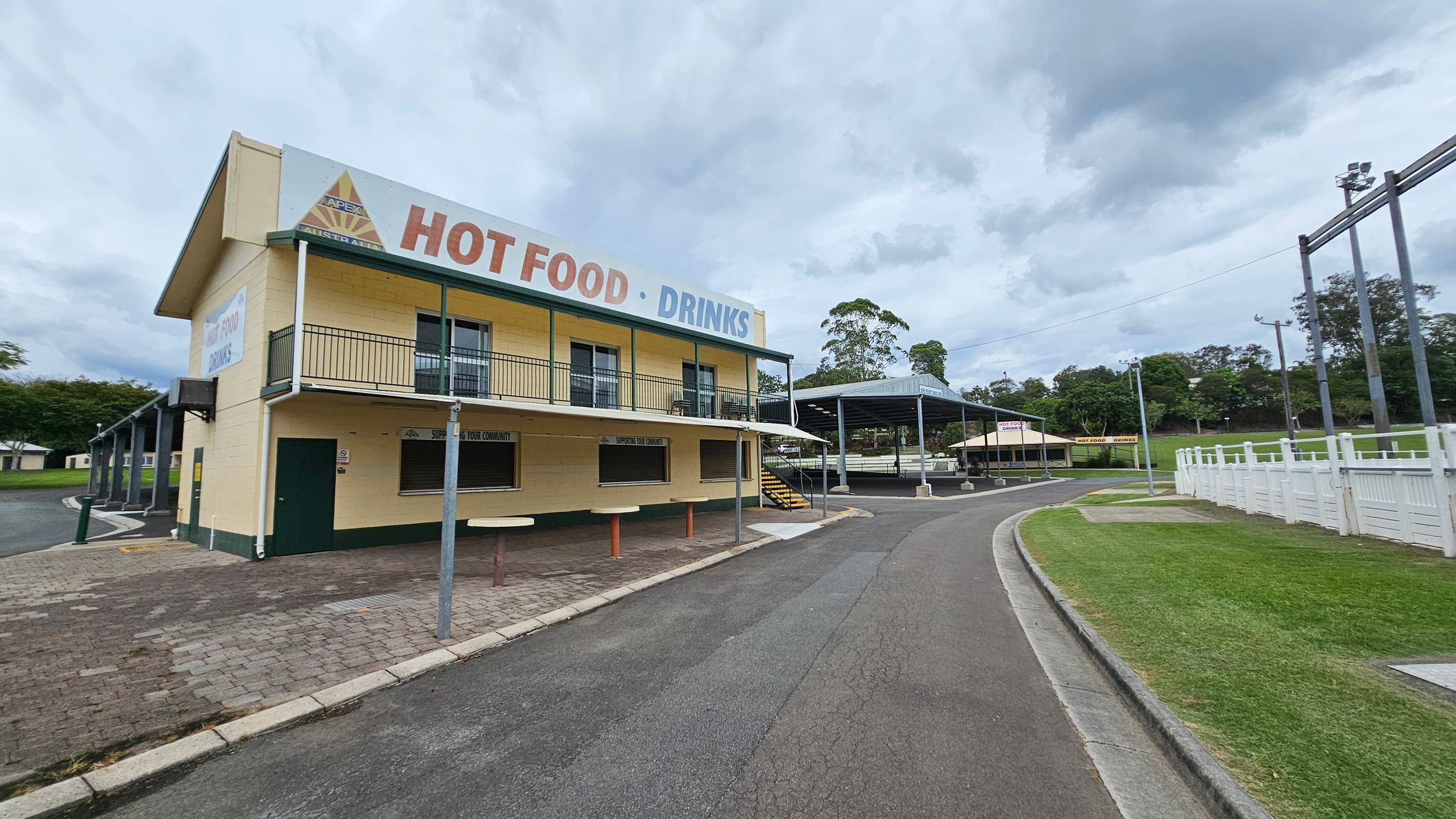 A two-storey yellow building with green trim on the left, featuring a sign that reads "HOT FOOD · DRINKS". It has a balcony and a service window on the ground floor. A paved path leads to a covered seating area with a metal roof in the centre. On the right, there's a curved road bordered by a white fence and a grassy area. Trees and additional structures are visible in the background. A two-storey yellow building with green trim on the left, featuring a sign that reads "HOT FOOD · DRINKS". It has a balcony and a service window on the ground floor. A paved path leads to a covered seating area with a metal roof in the centre. On the right, there's a curved road bordered by a white fence and a grassy area. Trees and additional structures are visible in the background.