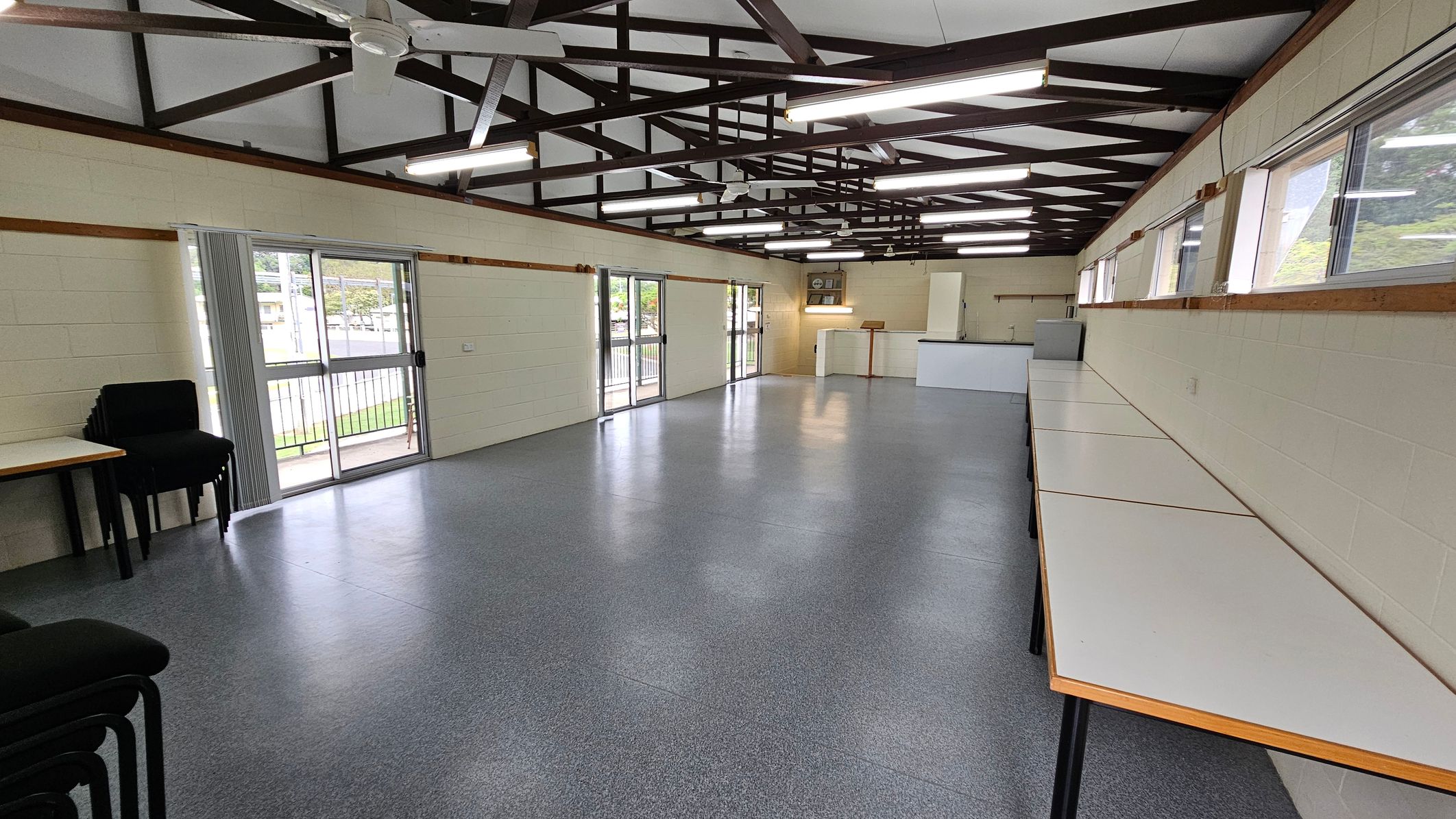 A spacious room with a grey speckled floor and a high ceiling featuring exposed beams and fluorescent lights. On the left, there are stacked black chairs and a sliding glass door leading outside. The right side has a long row of white tables against the wall with windows above. At the far end, there is a white counter and a small wooden lectern. A spacious room with a grey speckled floor and a high ceiling featuring exposed beams and fluorescent lights. On the left, there are stacked black chairs and a sliding glass door leading outside. The right side has a long row of white tables against the wall with windows above. At the far end, there is a white counter and a small wooden lectern.