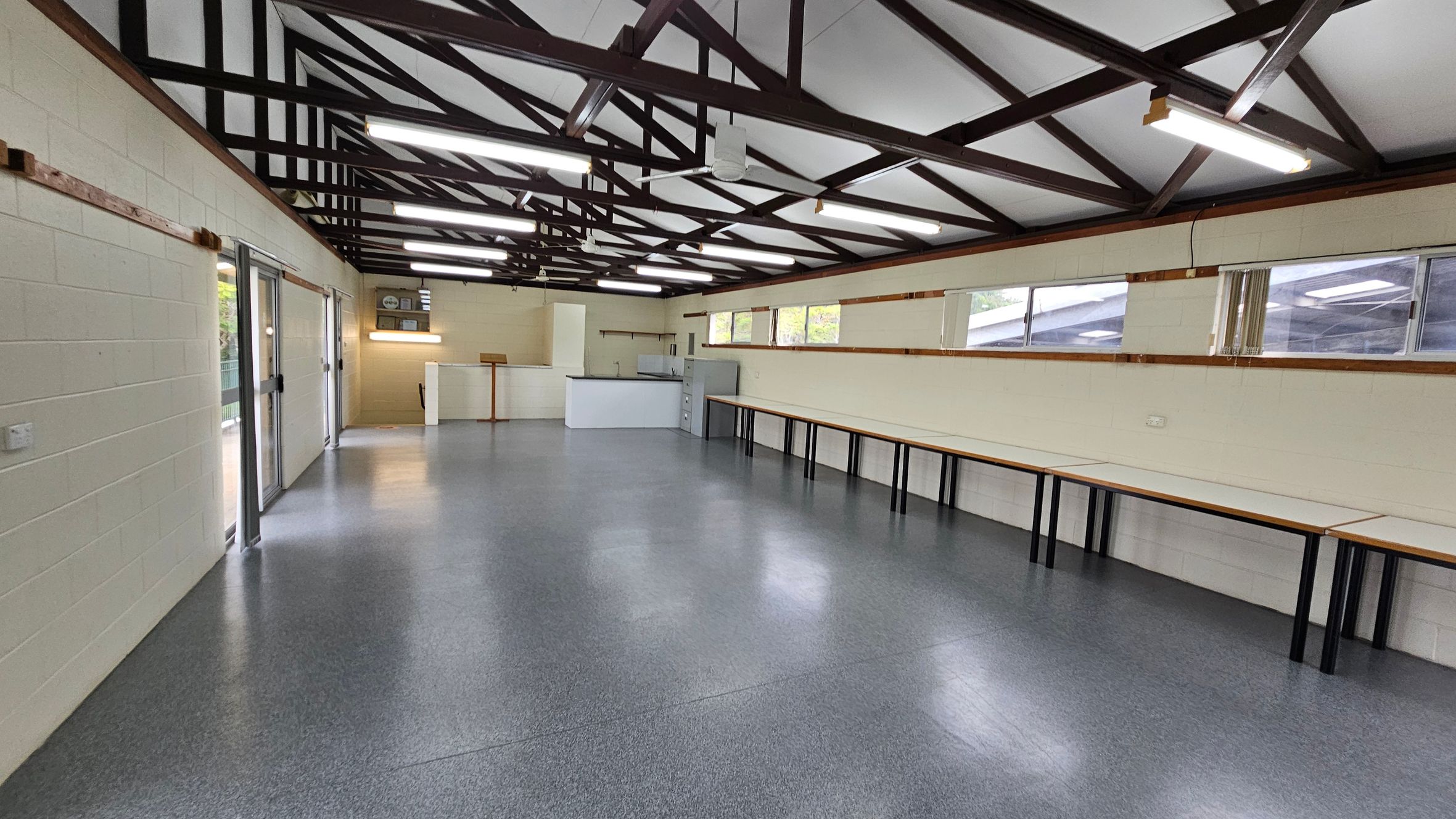 A spacious room with a grey speckled floor and a high ceiling featuring exposed beams. On the left, there are large glass doors. The right side has a long row of tables against the wall, beneath several windows. At the far end, there is a small counter area with a few cabinets. Fluorescent lights are mounted on the ceiling, providing bright illumination. A spacious room with a grey speckled floor and a high ceiling featuring exposed beams. On the left, there are large glass doors. The right side has a long row of tables against the wall, beneath several windows. At the far end, there is a small counter area with a few cabinets. Fluorescent lights are mounted on the ceiling, providing bright illumination.