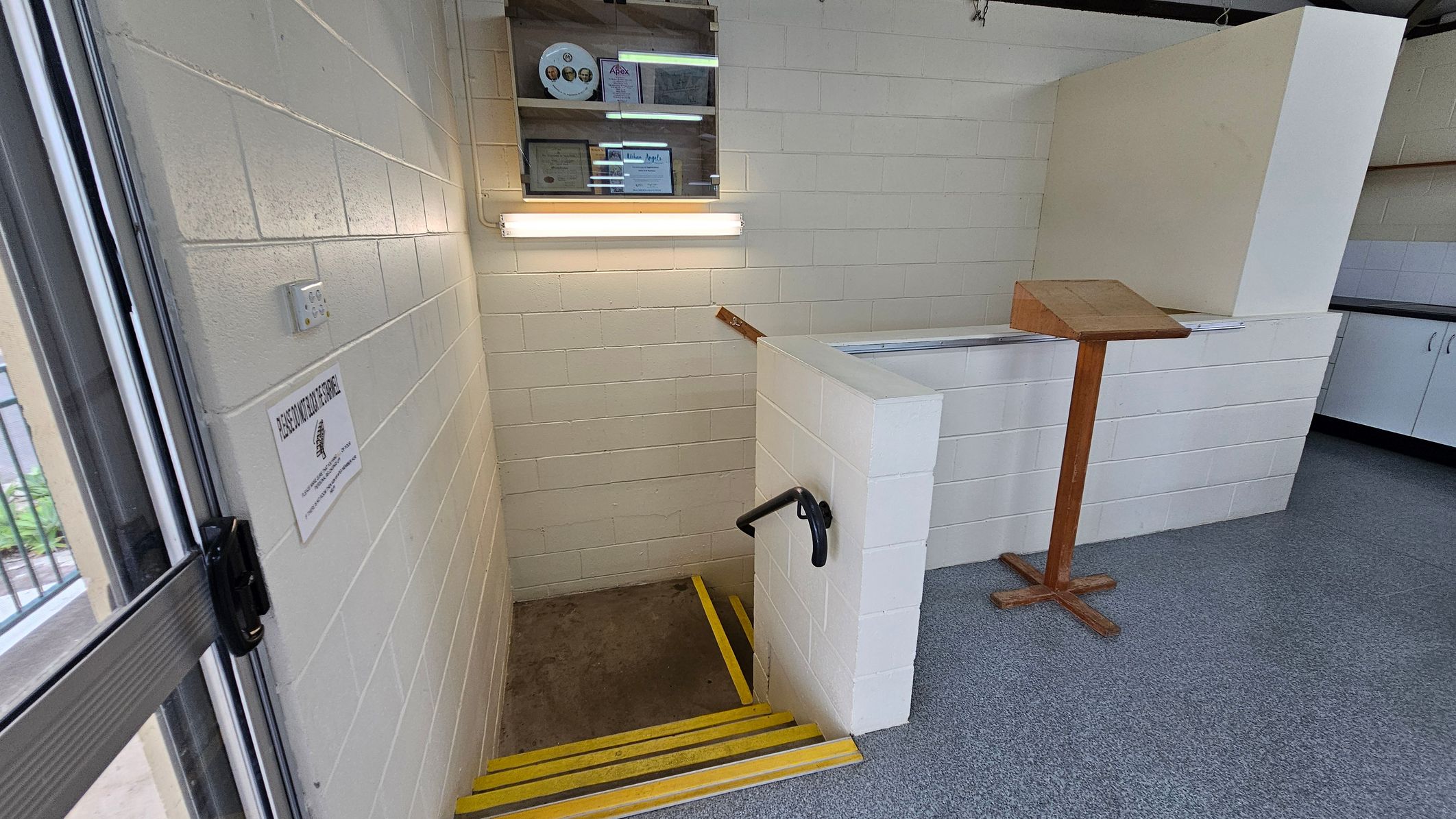 A small stairwell with yellow-edged steps descends into a lower level. On the left, a white brick wall features a sign and a light switch. Above the stairs, a wall-mounted display case holds certificates and a circular item. On the right, a wooden lectern stands on a grey carpeted floor. The ceiling is not visible. A small stairwell with yellow-edged steps descends into a lower level. On the left, a white brick wall features a sign and a light switch. Above the stairs, a wall-mounted display case holds certificates and a circular item. On the right, a wooden lectern stands on a grey carpeted floor. The ceiling is not visible.