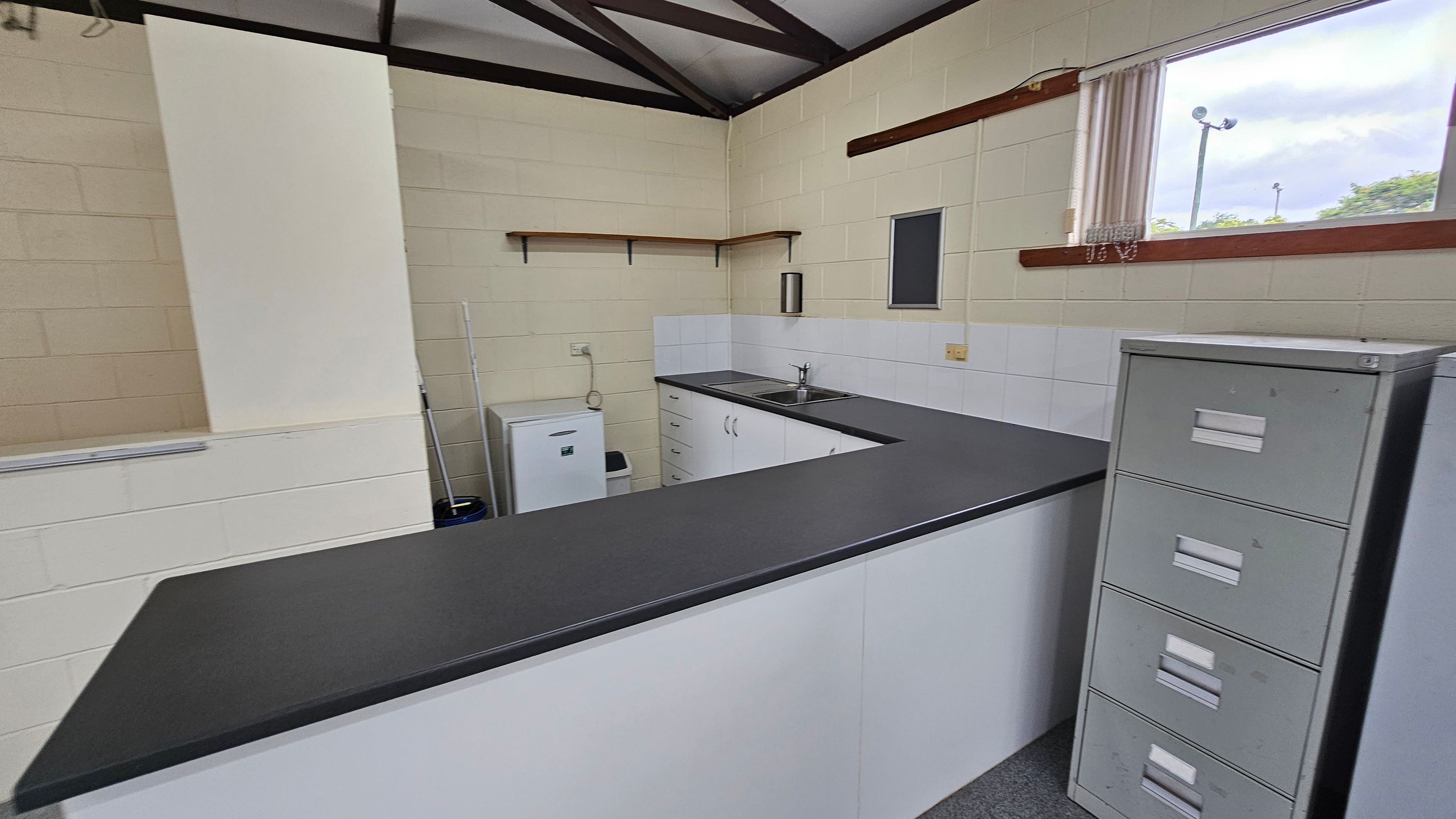 A small kitchen area with a U-shaped black countertop. On the left, there's a white mini fridge and a mop. The back wall has a sink with a double basin, white cabinets, and a small shelf above. On the right, a window with vertical blinds lets in natural light. A grey filing cabinet stands next to the counter. The walls are light green with white tiles around the sink area. The ceiling has exposed beams. A small kitchen area with a U-shaped black countertop. On the left, there's a white mini fridge and a mop. The back wall has a sink with a double basin, white cabinets, and a small shelf above. On the right, a window with vertical blinds lets in natural light. A grey filing cabinet stands next to the counter. The walls are light green with white tiles around the sink area. The ceiling has exposed beams.