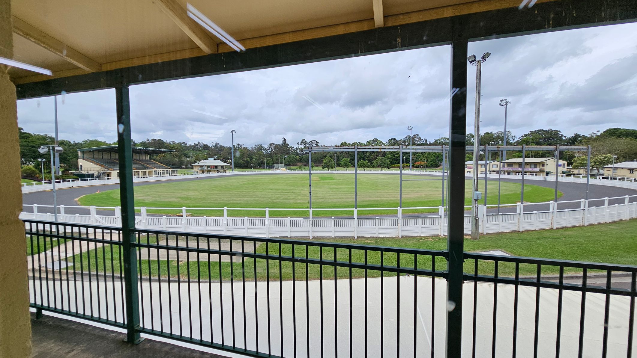 A view from a covered balcony overlooking a circular sports field. The foreground features a black metal railing. Beyond the railing, there's a grassy field encircled by a paved track and a white fence. In the background, there are several buildings, including a grandstand on the left and smaller structures on the right. Tall floodlights are positioned around the field, and trees are visible in the distance. The ceiling of the balcony is wooden with visible beams. A view from a covered balcony overlooking a circular sports field. The foreground features a black metal railing. Beyond the railing, there's a grassy field encircled by a paved track and a white fence. In the background, there are several buildings, including a grandstand on the left and smaller structures on the right. Tall floodlights are positioned around the field, and trees are visible in the distance. The ceiling of the balcony is wooden with visible beams.