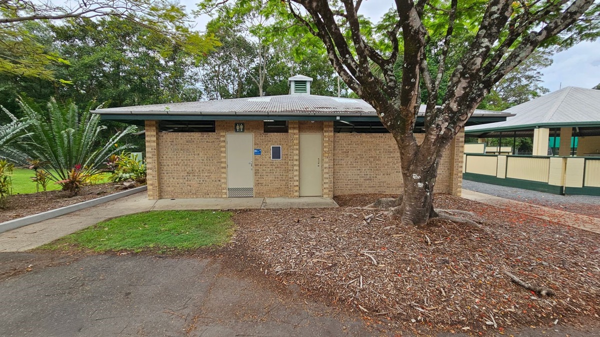 A single-storey brick building with two closed doors in the centre and right. On the left, a concrete path curves alongside a garden with green foliage and small shrubs. A large tree with an uneven bark trunk is in the foreground to the right, surrounded by woodchip ground cover. In the background, another structure with cream walls and a green roof is partially visible. The floor consists of concrete, tarmac, and soil. The ceiling of the visible building is metal roofing. A single-storey brick building with two closed doors in the centre and right. On the left, a concrete path curves alongside a garden with green foliage and small shrubs. A large tree with an uneven bark trunk is in the foreground to the right, surrounded by woodchip ground cover. In the background, another structure with cream walls and a green roof is partially visible. The floor consists of concrete, tarmac, and soil. The ceiling of the visible building is metal roofing.