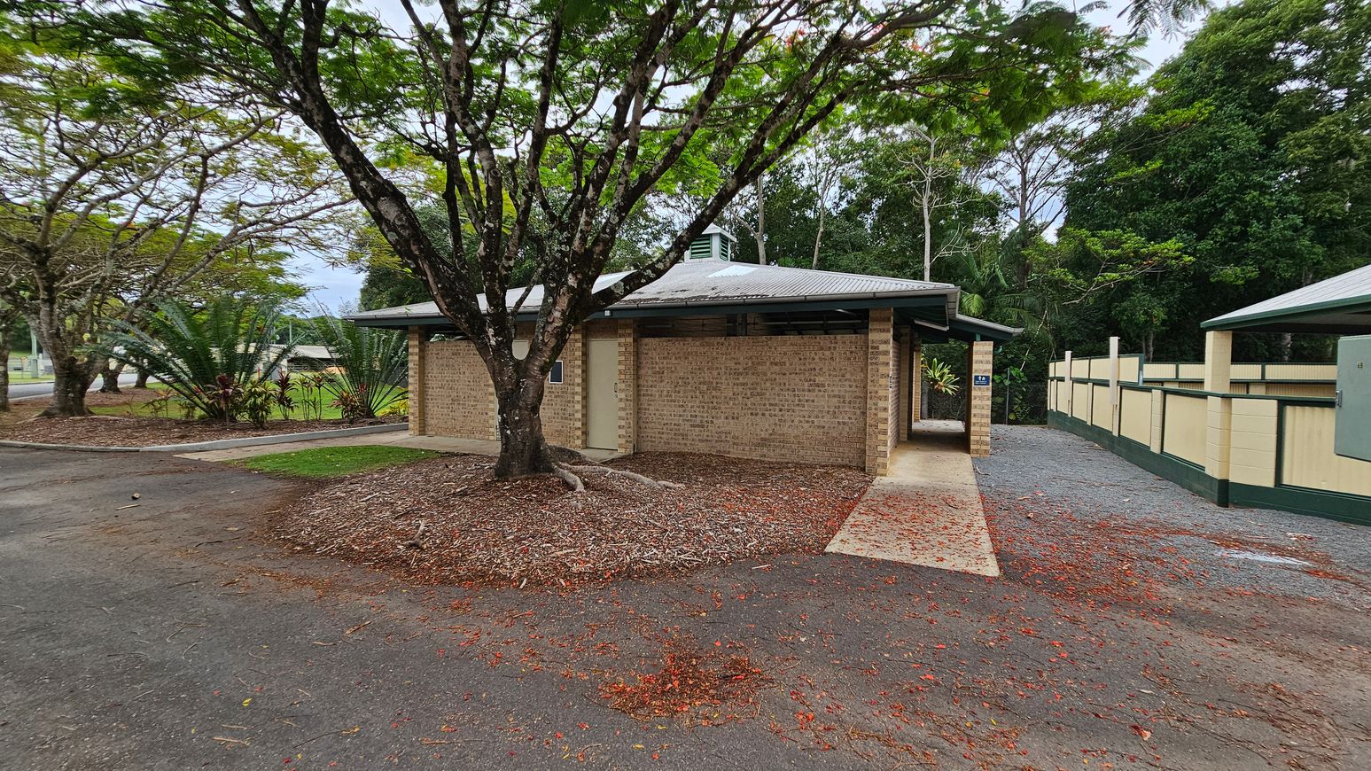 A brick building stands in the centre, with a metal roof and an open passage on the right. On the left, a large tree with a wide, textured trunk is surrounded by woodchip ground cover. On the right, a paved walkway runs alongside a cream and green fence. The ground is a mix of tarmac, concrete, and scattered red leaves. In the background, tall trees and foliage create a dense green backdrop. A brick building stands in the centre, with a metal roof and an open passage on the right. On the left, a large tree with a wide, textured trunk is surrounded by woodchip ground cover. On the right, a paved walkway runs alongside a cream and green fence. The ground is a mix of tarmac, concrete, and scattered red leaves. In the background, tall trees and foliage create a dense green backdrop.