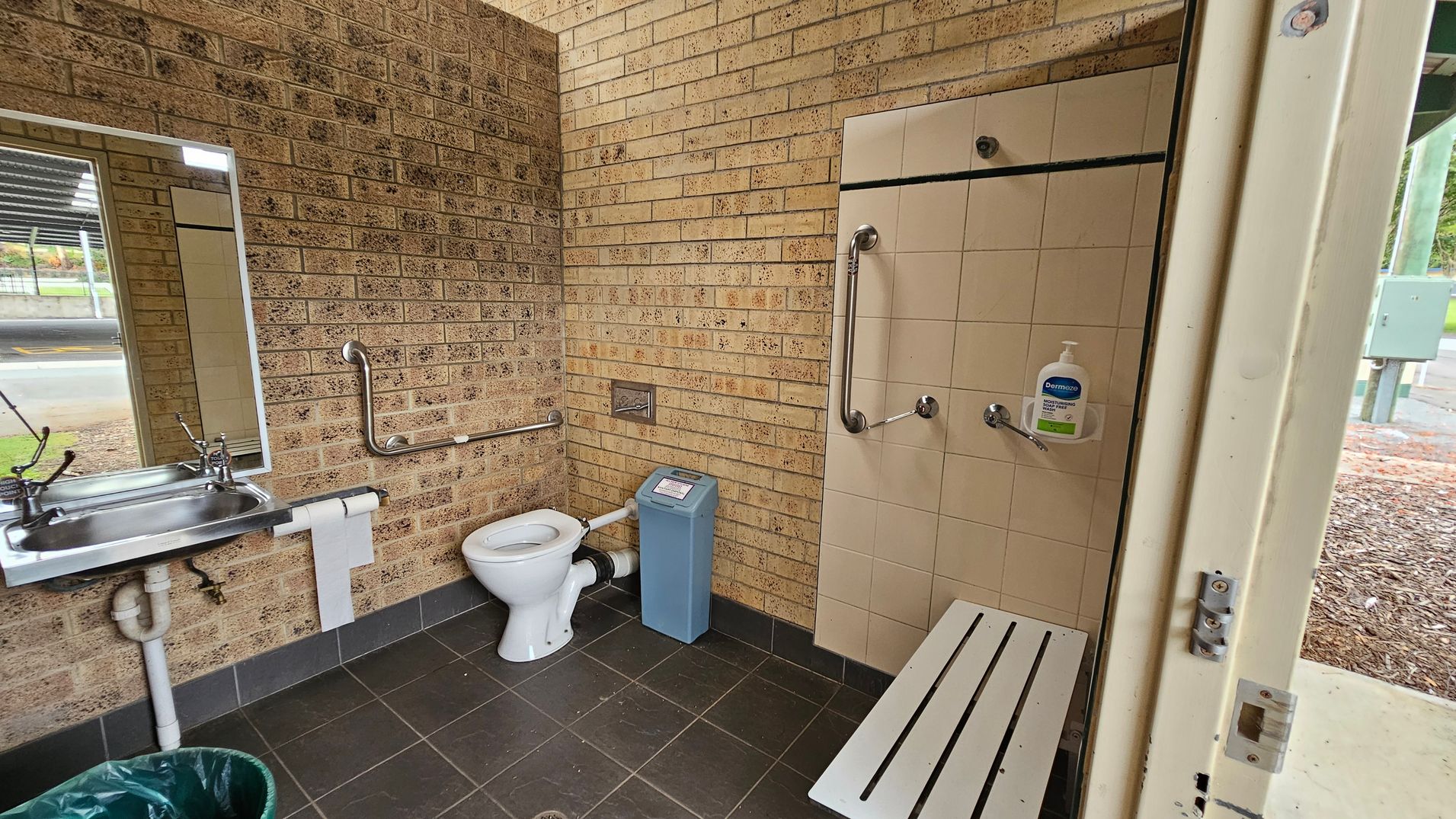 A public restroom with beige brick walls and dark tiled flooring. On the left, a stainless steel sink with a mirror above it and a mounted toilet roll holder beneath. In the middle, a white toilet with a grab bar on the wall behind and a blue sanitary bin to the right. On the right, a tiled wall with attached grab bars, a bottle of soap, and a white slatted bench extending below. The ceiling is cream-coloured with visible piping. A public restroom with beige brick walls and dark tiled flooring. On the left, a stainless steel sink with a mirror above it and a mounted toilet roll holder beneath. In the middle, a white toilet with a grab bar on the wall behind and a blue sanitary bin to the right. On the right, a tiled wall with attached grab bars, a bottle of soap, and a white slatted bench extending below. The ceiling is cream-coloured with visible piping.