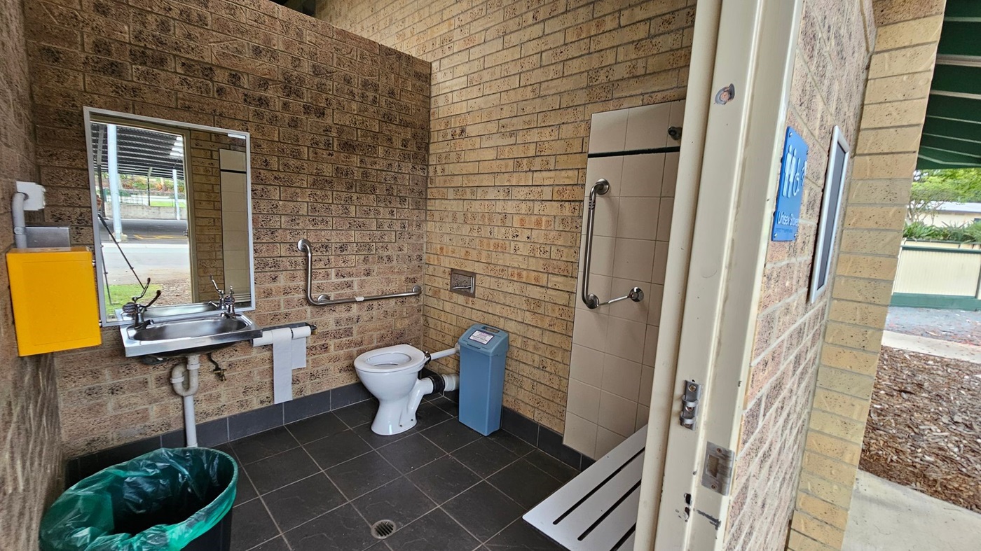 A disabled-access toilet with tan brick walls and dark grey tiled flooring. On the left, a stainless steel sink with a mirror above, a yellow soap dispenser, and a green bin below. In the middle, a white toilet with metal grab rails and a blue sanitary bin beside it. On the right, a tiled shower area with grab bars and a horizontal white bench. The ceiling is light-coloured with exposed piping. The doorway on the right opens to an outdoor area with bark mulch and a green-roofed structure. A disabled-access toilet with tan brick walls and dark grey tiled flooring. On the left, a stainless steel sink with a mirror above, a yellow soap dispenser, and a green bin below. In the middle, a white toilet with metal grab rails and a blue sanitary bin beside it. On the right, a tiled shower area with grab bars and a horizontal white bench. The ceiling is light-coloured with exposed piping. The doorway on the right opens to an outdoor area with bark mulch and a green-roofed structure.