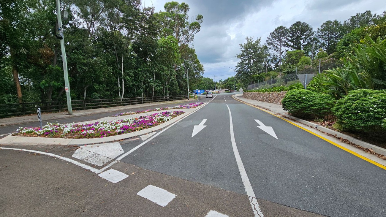 A road curves to the left with white directional arrows painted on it. On the right, a large tree with sprawling roots stands next to a blue sign that reads "Welcome to Nambour Showgrounds." In the background, there is a small building with a light-coloured exterior and a gate. The area is surrounded by lush greenery and trees. The ground is covered with fallen leaves. A road curves to the left with white directional arrows painted on it. On the right, a large tree with sprawling roots stands next to a blue sign that reads "Welcome to Nambour Showgrounds." In the background, there is a small building with a light-coloured exterior and a gate. The area is surrounded by lush greenery and trees. The ground is covered with fallen leaves.