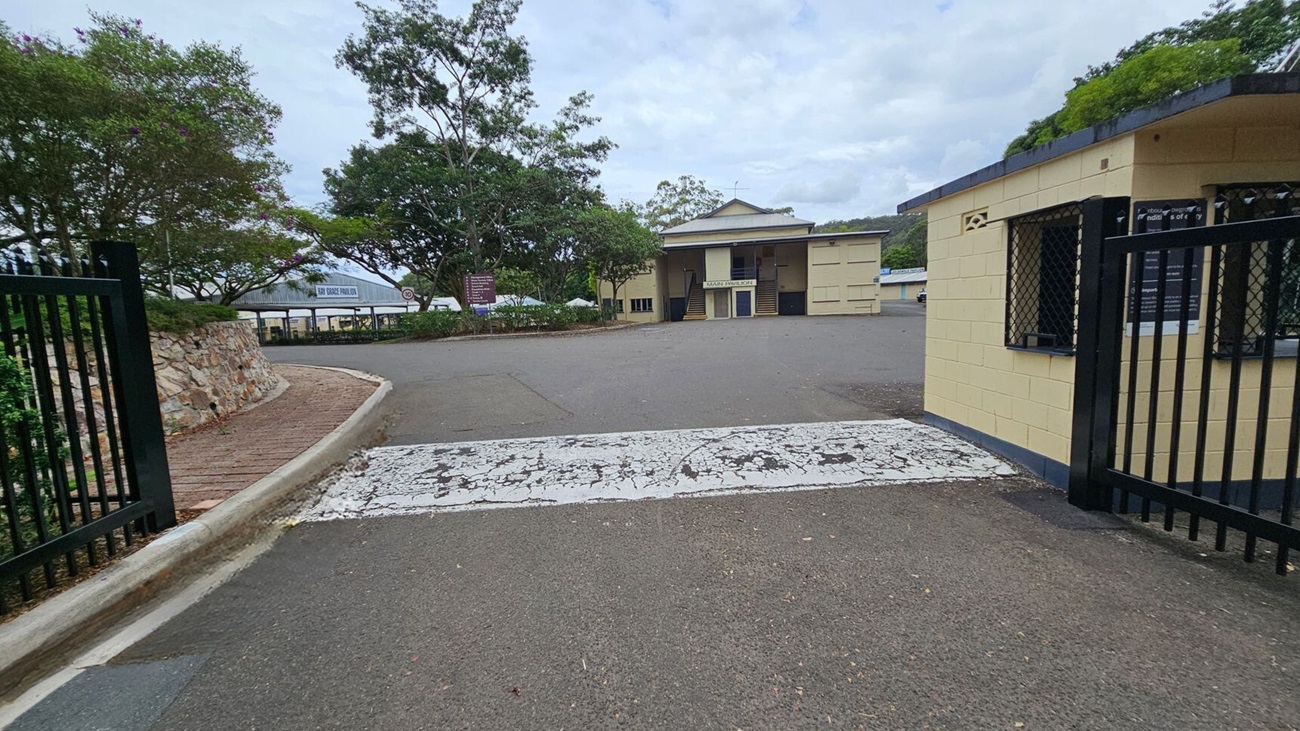 A paved road leads to a building with a sign reading "Main Pavilion" in the centre. On the left, there's a stone wall with trees and a path curving alongside it. On the right, a small yellow building with barred windows is adjacent to a black metal gate. The ground is marked with a worn white pedestrian crossing. Trees and additional structures are visible in the background. A paved road leads to a building with a sign reading "Main Pavilion" in the centre. On the left, there's a stone wall with trees and a path curving alongside it. On the right, a small yellow building with barred windows is adjacent to a black metal gate. The ground is marked with a worn white pedestrian crossing. Trees and additional structures are visible in the background.