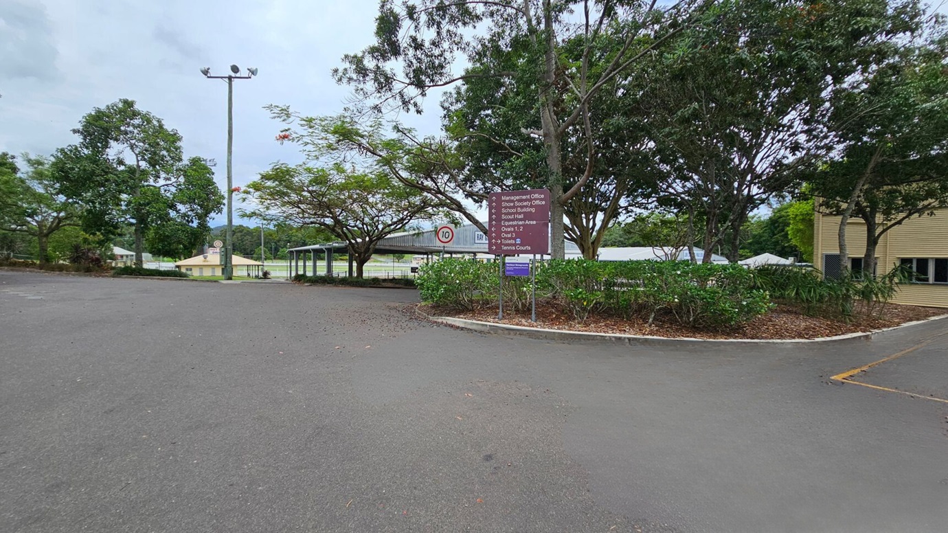 A two-storey building labelled "Main Pavilion" is in the centre, with a staircase leading to the entrance. The building is light-coloured with a gabled roof. On the left, there are trees and shrubs lining a path. On the right, more dense trees and several green bins are visible. The foreground features a large, empty paved area with some scattered leaves and a marked parking space. A two-storey building labelled "Main Pavilion" is in the centre, with a staircase leading to the entrance. The building is light-coloured with a gabled roof. On the left, there are trees and shrubs lining a path. On the right, more dense trees and several green bins are visible. The foreground features a large, empty paved area with some scattered leaves and a marked parking space.