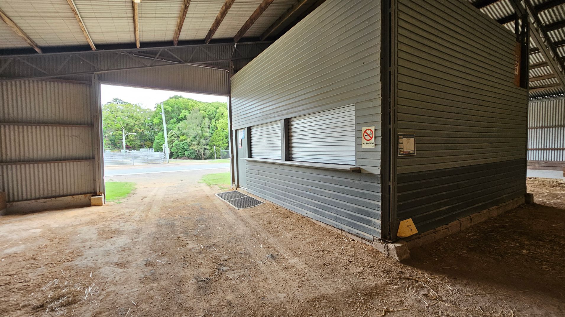 A large grey metal building labelled "Fergus Scott Pavilion" and "Dairy Cattle" is in the centre. It has two large open entrances. To the left, there's a smaller grey shed. The ground in front is a mix of gravel and grass. On the right, part of another structure with a sloped roof is visible. Trees are in the background on the left. A large grey metal building labelled "Fergus Scott Pavilion" and "Dairy Cattle" is in the centre. It has two large open entrances. To the left, there's a smaller grey shed. The ground in front is a mix of gravel and grass. On the right, part of another structure with a sloped roof is visible. Trees are in the background on the left.