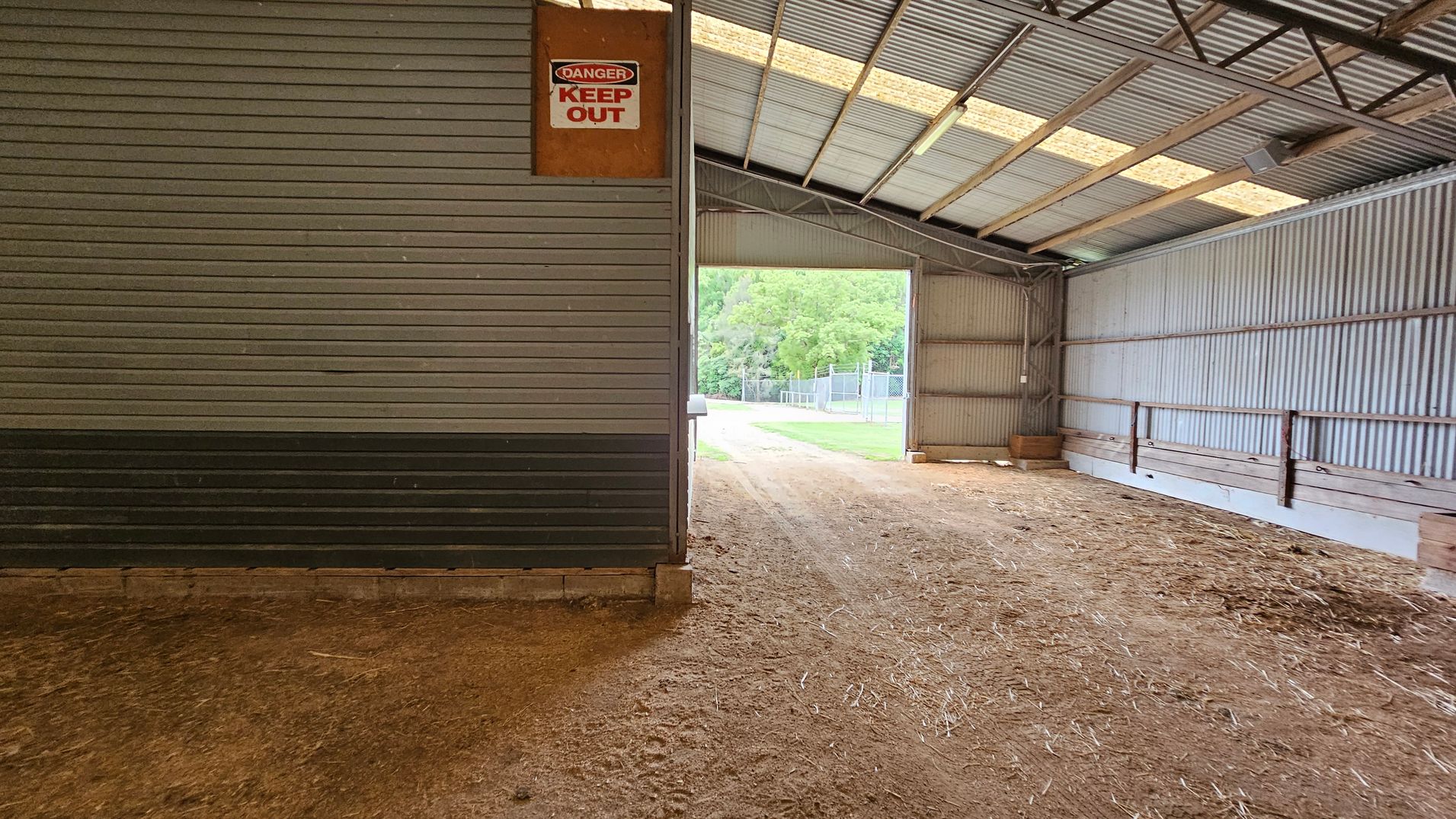 Inside a large, open-sided shed with a corrugated metal roof and walls. On the right, there's a small structure with horizontal cladding and closed shutters, featuring a "No Smoking" sign. The floor is dirt, with some grass and debris. Outside, visible through the open side, is a road bordered by green trees and a metal fence. The ceiling is high with exposed beams. Inside a large, open-sided shed with a corrugated metal roof and walls. On the right, there's a small structure with horizontal cladding and closed shutters, featuring a "No Smoking" sign. The floor is dirt, with some grass and debris. Outside, visible through the open side, is a road bordered by green trees and a metal fence. The ceiling is high with exposed beams.