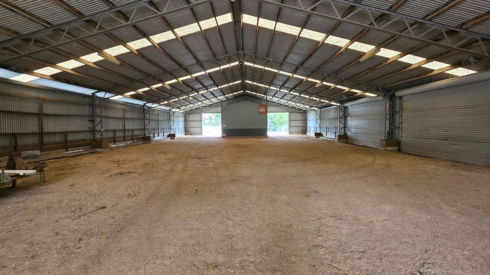 A large, empty warehouse with a corrugated metal roof and walls. The floor is covered with a mix of dirt and gravel. The ceiling has translucent panels allowing natural light to filter in. On the far end, there are two large open doorways with views of greenery outside. The interior is supported by metal beams, and there are some wooden pallets and equipment on the left side. The space is expansive and industrial in appearance. A large, empty warehouse with a corrugated metal roof and walls. The floor is covered with a mix of dirt and gravel. The ceiling has translucent panels allowing natural light to filter in. On the far end, there are two large open doorways with views of greenery outside. The interior is supported by metal beams, and there are some wooden pallets and equipment on the left side. The space is expansive and industrial in appearance.