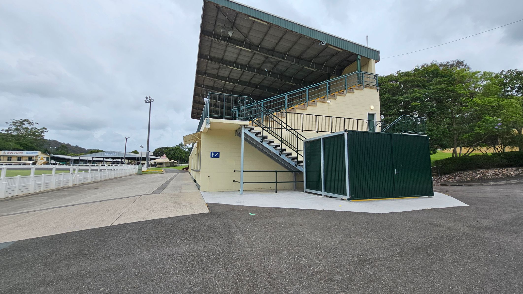 A sports facility with a covered grandstand on the right, featuring a row of green doors and large windows. The building is cream-coloured with green accents. The foreground shows a wide concrete pathway with patches of moss or lichen. In the background, there are tall floodlights and trees, with a smaller building visible to the left. The area appears open and spacious. A sports facility with a covered grandstand on the right, featuring a row of green doors and large windows. The building is cream-coloured with green accents. The foreground shows a wide concrete pathway with patches of moss or lichen. In the background, there are tall floodlights and trees, with a smaller building visible to the left. The area appears open and spacious.