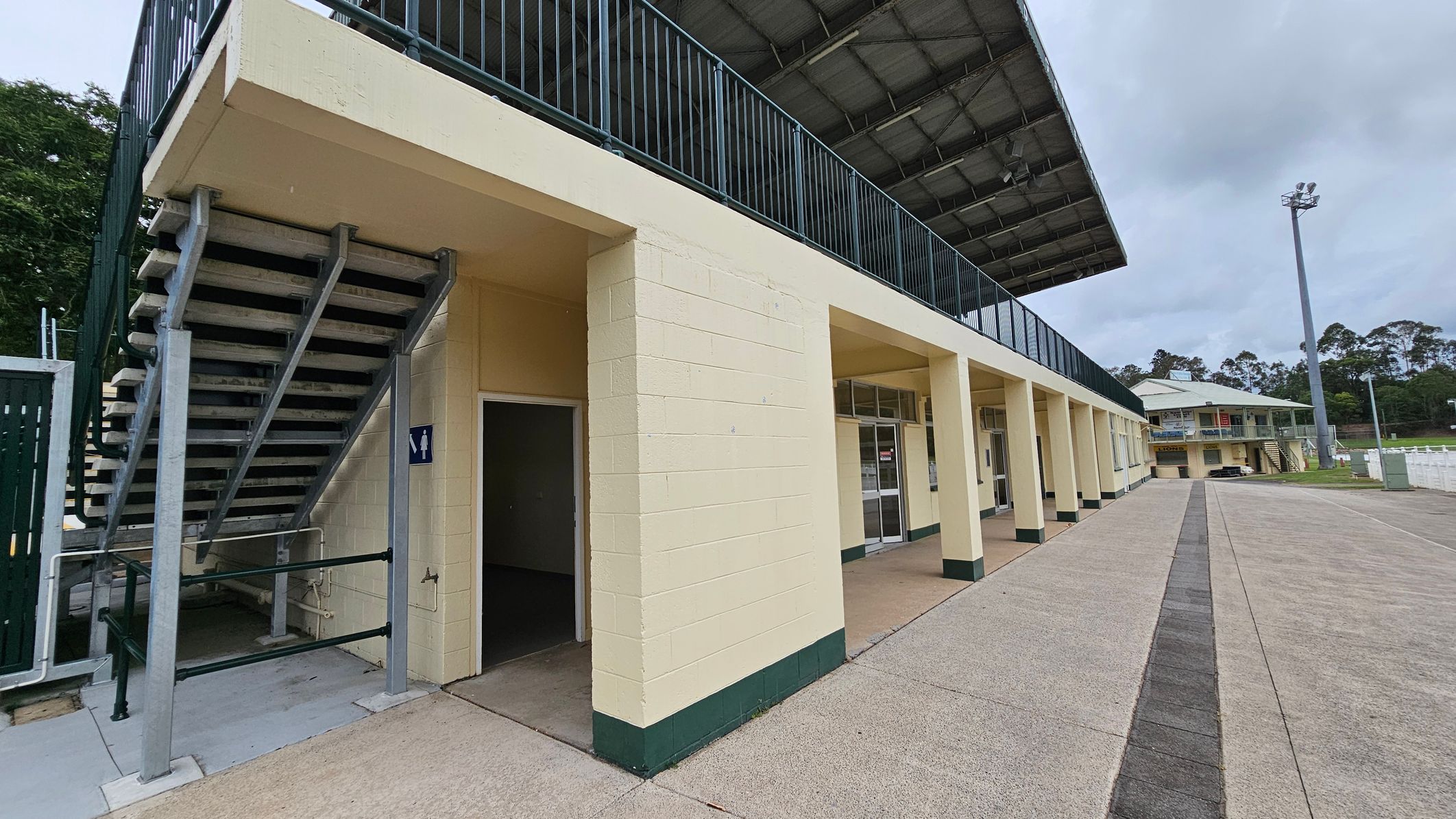 A sports facility with a two-storey structure. On the left, there is a staircase leading to the upper level, with a sign indicating a restroom below. The building has a series of columns and glass doors along the ground floor. The upper level features a railing and a covered roof. In the background, there is another similar building and a tall floodlight. The ground is paved with concrete, and there are trees in the distance. A sports facility with a two-storey structure. On the left, there is a staircase leading to the upper level, with a sign indicating a restroom below. The building has a series of columns and glass doors along the ground floor. The upper level features a railing and a covered roof. In the background, there is another similar building and a tall floodlight. The ground is paved with concrete, and there are trees in the distance.