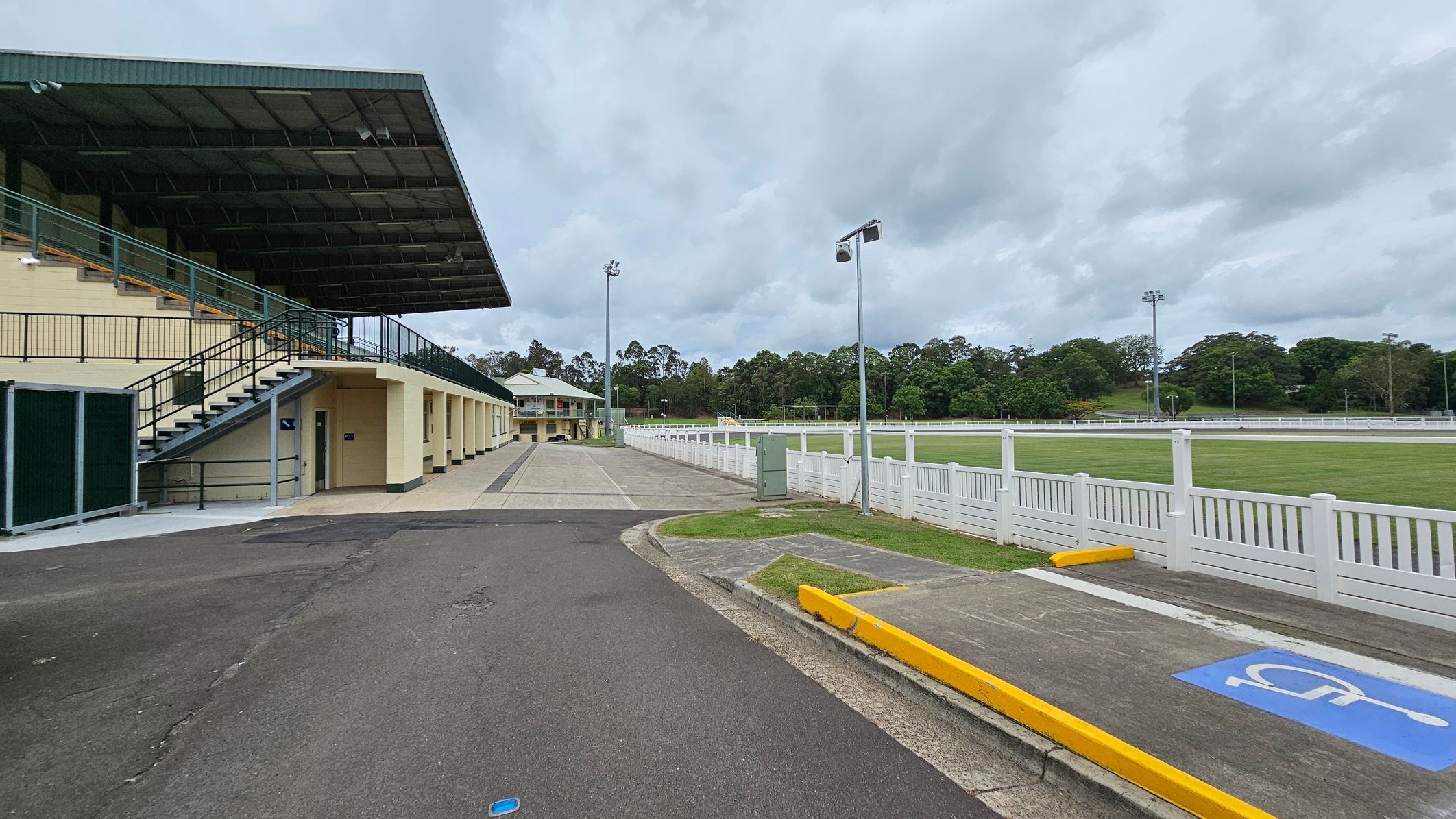 A sports facility with a covered grandstand on the left, featuring a staircase and multiple entrances. The ground is paved with a mix of asphalt and concrete. On the right, a white fence borders a grassy field. In the background, there are tall floodlights and a line of trees. A marked disabled parking space with a yellow kerb is visible in the foreground. A sports facility with a covered grandstand on the left, featuring a staircase and multiple entrances. The ground is paved with a mix of asphalt and concrete. On the right, a white fence borders a grassy field. In the background, there are tall floodlights and a line of trees. A marked disabled parking space with a yellow kerb is visible in the foreground.