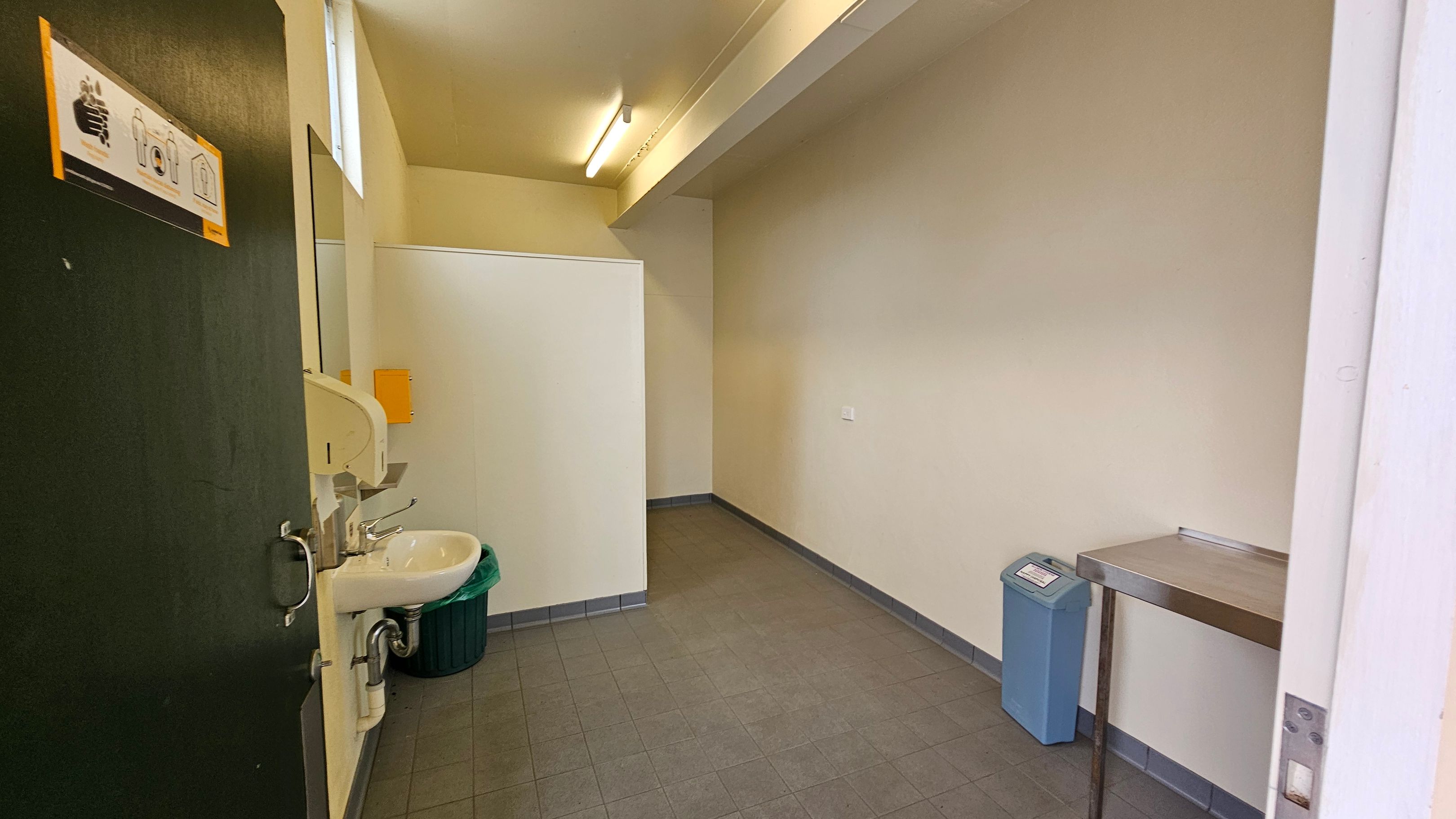 A small room with a tiled floor and a light-coloured ceiling. On the left, there's a partially open door with a handwashing instruction sign. Next to it is a white sink with a soap dispenser and a green bin underneath. On the right, a metal table is attached to the wall, and a blue bin is nearby. The back wall is plain, with a small section of a partition visible. The room is lit by a fluorescent light. A small room with a tiled floor and a light-coloured ceiling. On the left, there's a partially open door with a handwashing instruction sign. Next to it is a white sink with a soap dispenser and a green bin underneath. On the right, a metal table is attached to the wall, and a blue bin is nearby. The back wall is plain, with a small section of a partition visible. The room is lit by a fluorescent light.