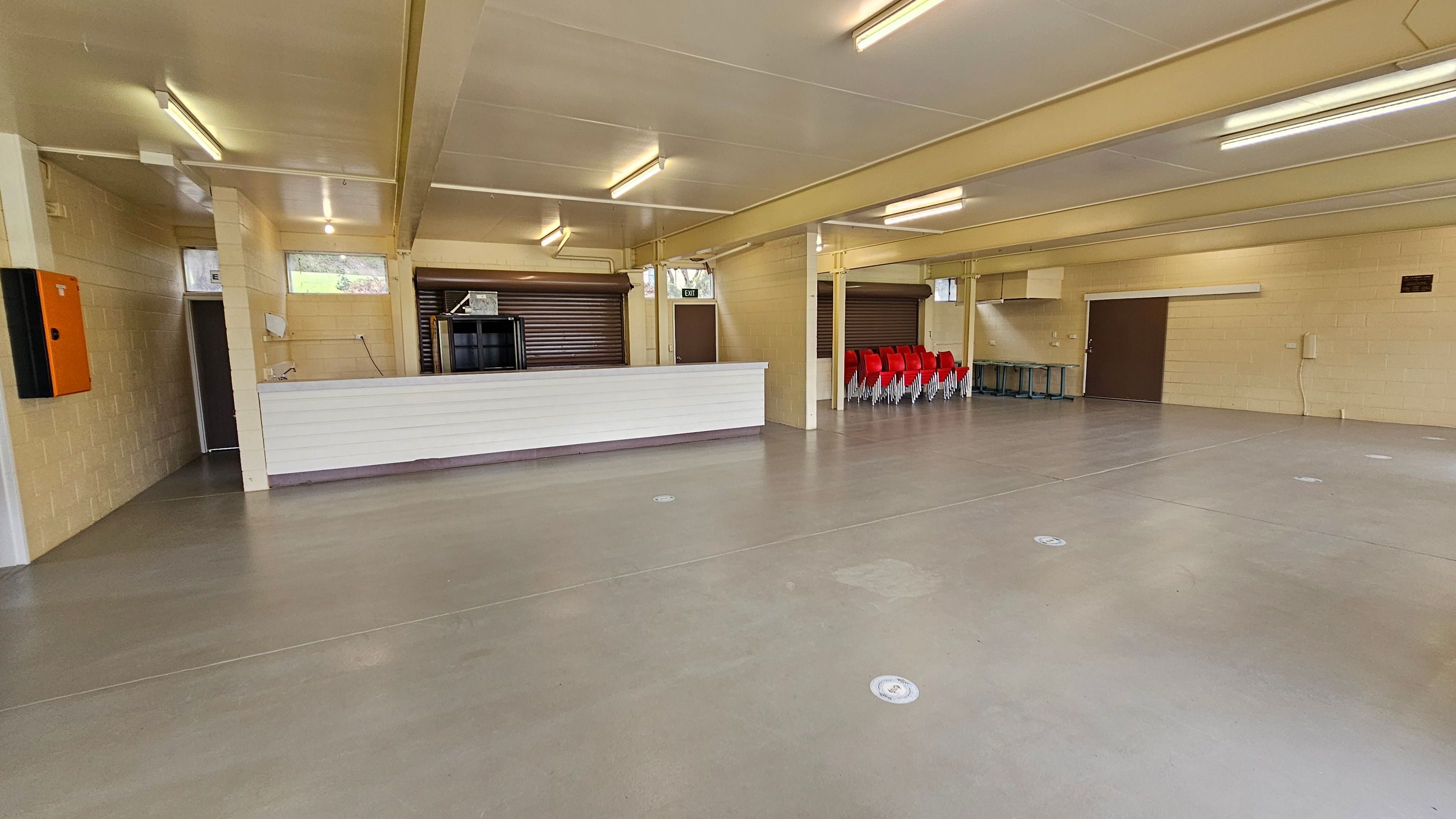 A spacious indoor area with a smooth, light brown floor and a white ceiling with fluorescent lights. On the left, there's a white counter with a small sink and an orange wall-mounted box. Behind the counter are two closed roller shutters. On the right, a stack of red chairs is neatly arranged against the wall. There are several doors, including an exit door, and a few small windows near the ceiling. The walls are light-coloured brick. A spacious indoor area with a smooth, light brown floor and a white ceiling with fluorescent lights. On the left, there's a white counter with a small sink and an orange wall-mounted box. Behind the counter are two closed roller shutters. On the right, a stack of red chairs is neatly arranged against the wall. There are several doors, including an exit door, and a few small windows near the ceiling. The walls are light-coloured brick.