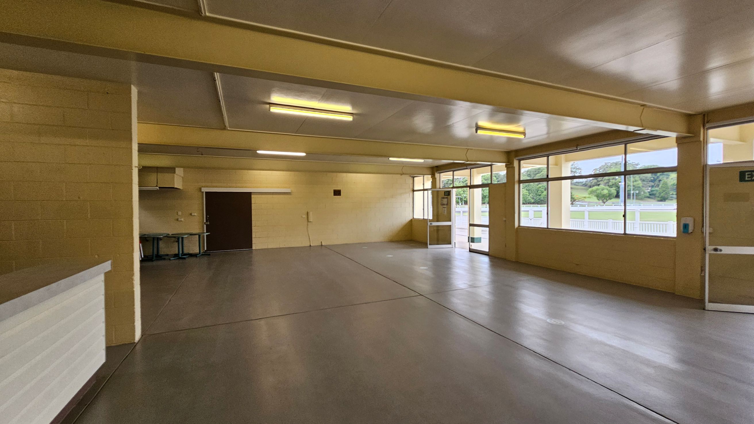 A spacious indoor room with a beige ceiling and floor. On the left, there's a white counter and a stack of red chairs against the wall. The back wall has a closed brown shutter and a door. On the right, large windows and a glass door provide natural light, with a view of an outdoor area. The room is lit by fluorescent lights on the ceiling. A spacious indoor room with a beige ceiling and floor. On the left, there's a white counter and a stack of red chairs against the wall. The back wall has a closed brown shutter and a door. On the right, large windows and a glass door provide natural light, with a view of an outdoor area. The room is lit by fluorescent lights on the ceiling.