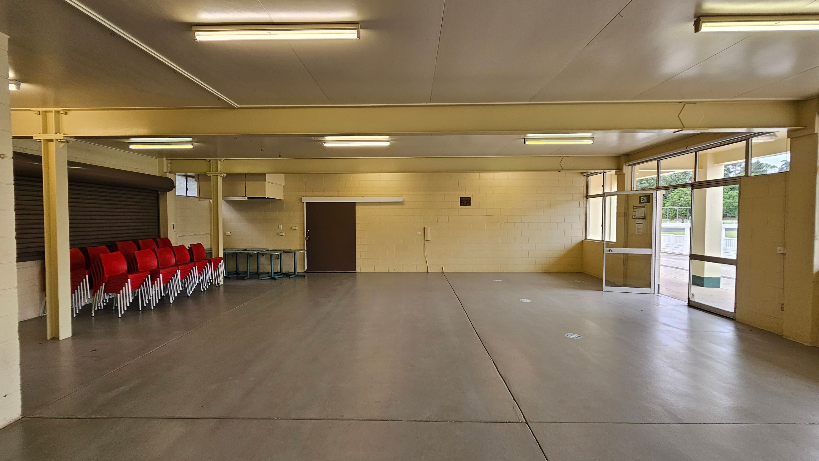 A spacious, empty room with beige walls and a smooth grey floor. On the left, there's a counter with a white top. The back wall features a dark brown door and a small kitchen area with a range hood. Large windows on the right let in natural light, showing a view of greenery outside. The ceiling is white with fluorescent lights. An exit door is on the far right. A spacious, empty room with beige walls and a smooth grey floor. On the left, there's a counter with a white top. The back wall features a dark brown door and a small kitchen area with a range hood. Large windows on the right let in natural light, showing a view of greenery outside. The ceiling is white with fluorescent lights. An exit door is on the far right.