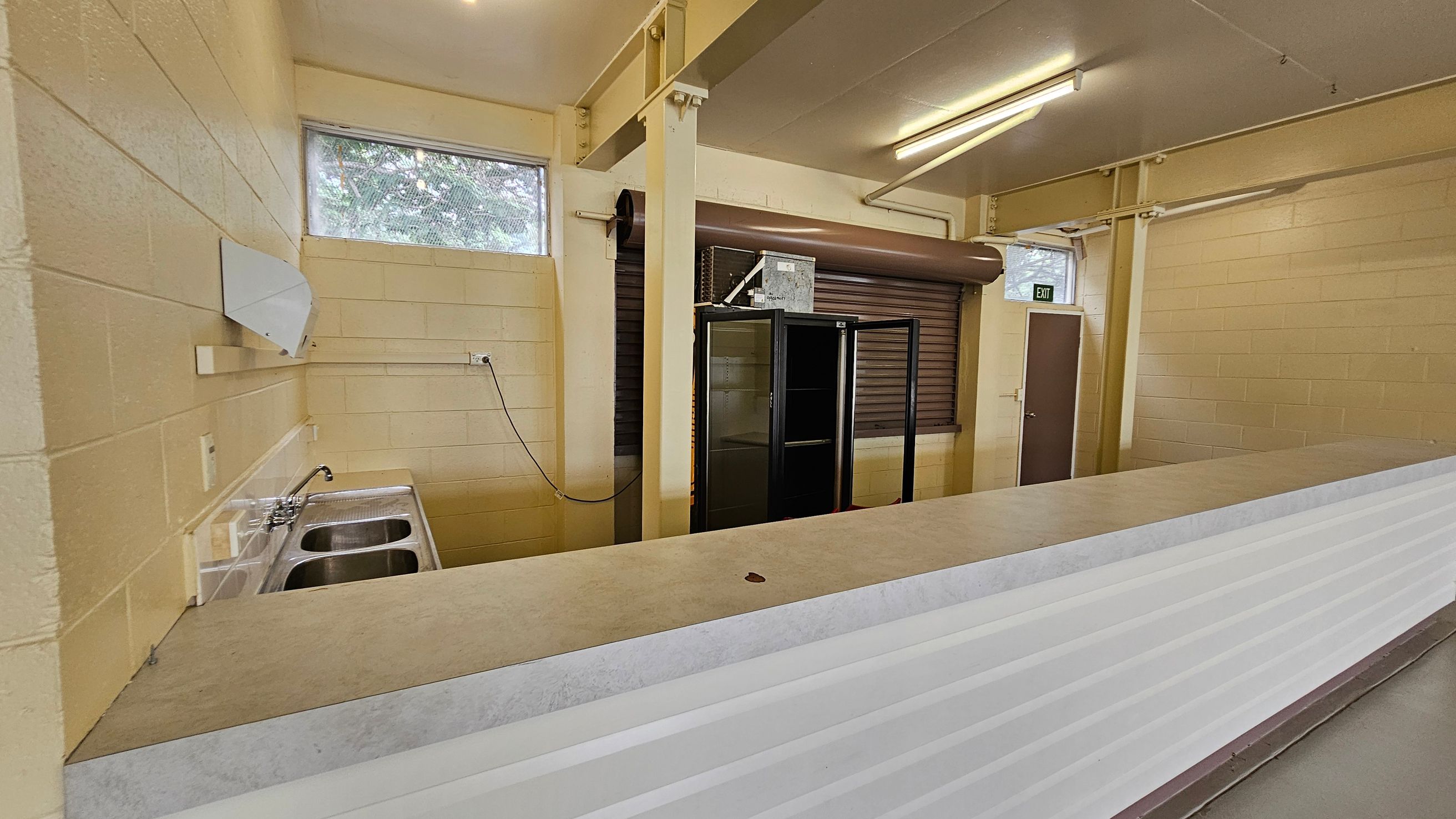 A room with cream-coloured brick walls and a long counter in the foreground. On the left, there's a double sink with a tap and a small window above it. In the middle, a black-framed glass cabinet stands next to a closed roller shutter. On the right, a brown door with an "EXIT" sign above it. The ceiling is fitted with fluorescent lights, and the floor is not visible. A room with cream-coloured brick walls and a long counter in the foreground. On the left, there's a double sink with a tap and a small window above it. In the middle, a black-framed glass cabinet stands next to a closed roller shutter. On the right, a brown door with an "EXIT" sign above it. The ceiling is fitted with fluorescent lights, and the floor is not visible.