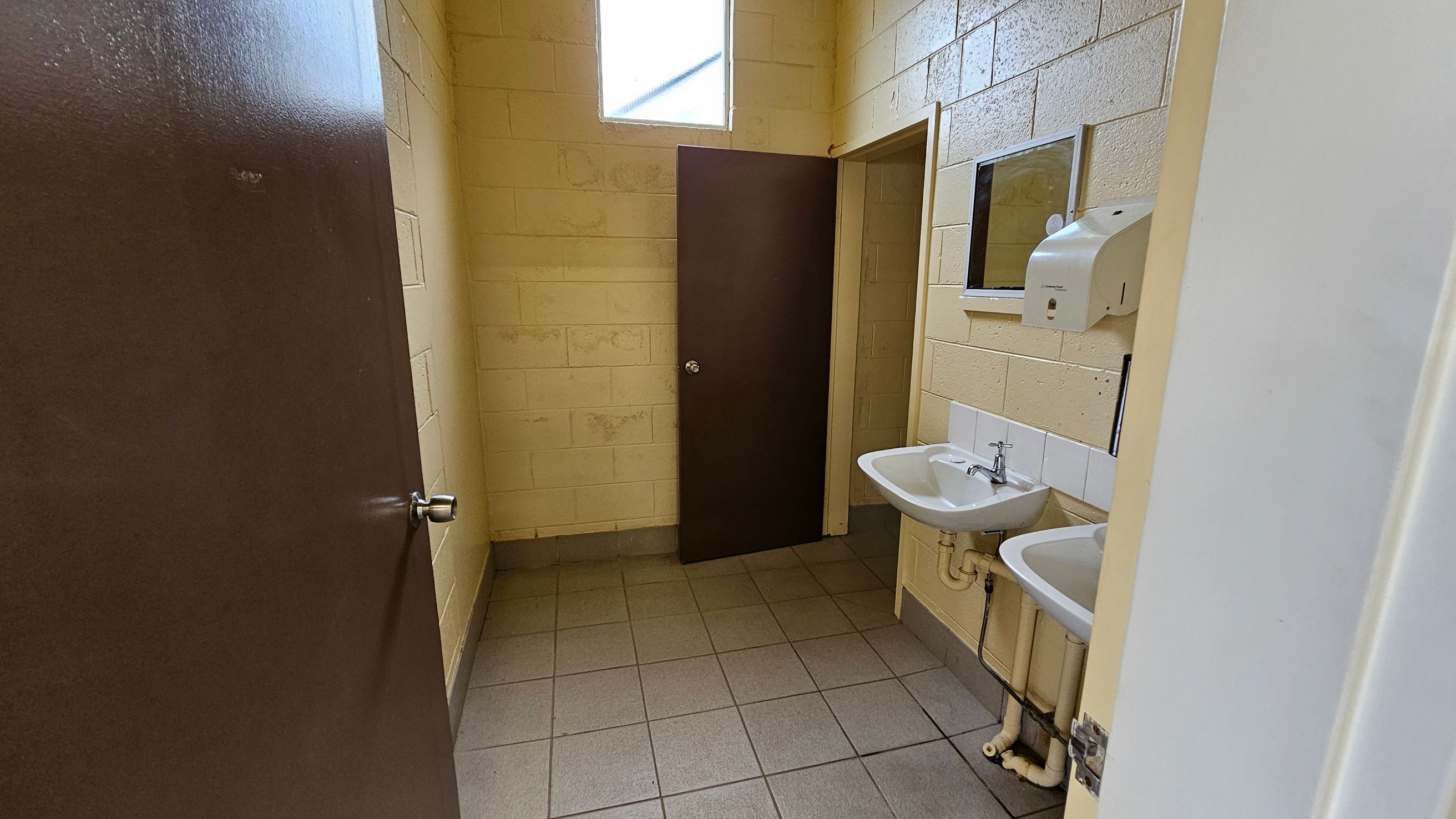 A small bathroom with beige brick walls and a tiled floor. On the right, there are two white sinks with a mirror above and a soap dispenser mounted on the wall. In the centre, a brown door is slightly ajar, leading to another room. Above the door, a high window allows natural light to enter. The ceiling is not visible. A small bathroom with beige brick walls and a tiled floor. On the right, there are two white sinks with a mirror above and a soap dispenser mounted on the wall. In the centre, a brown door is slightly ajar, leading to another room. Above the door, a high window allows natural light to enter. The ceiling is not visible.