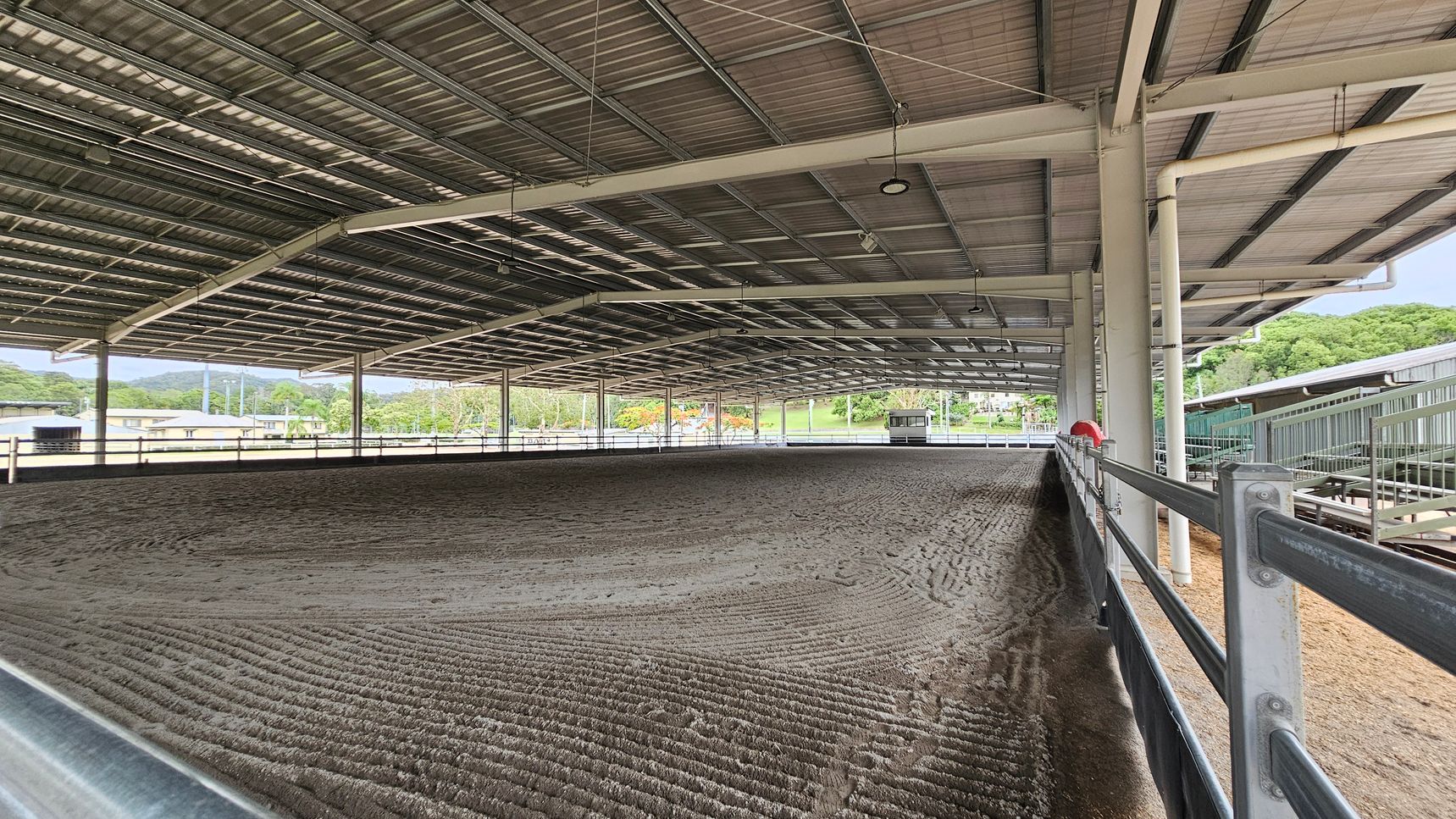 A large covered equestrian arena with a metal roof and open sides. The ground is textured with a mix of sand and soil. On the right, a metal railing runs along the perimeter. In the background, there are trees and some buildings visible through the open sides. The ceiling is supported by metal beams and has several lights hanging from it. A large covered equestrian arena with a metal roof and open sides. The ground is textured with a mix of sand and soil. On the right, a metal railing runs along the perimeter. In the background, there are trees and some buildings visible through the open sides. The ceiling is supported by metal beams and has several lights hanging from it.