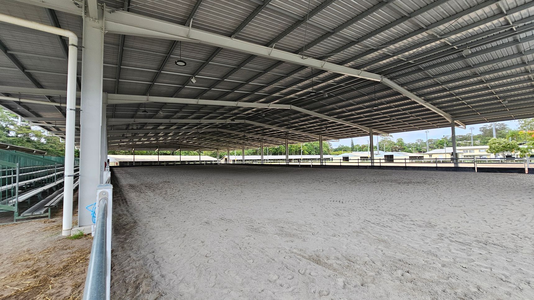A large covered equestrian arena with a metal roof and open sides. The ground is sandy, showing hoof prints. On the left, there are green metal bleachers for spectators. The background features trees and some buildings visible through the open sides. The ceiling is supported by metal beams and has several lights hanging from it. A large covered equestrian arena with a metal roof and open sides. The ground is sandy, showing hoof prints. On the left, there are green metal bleachers for spectators. The background features trees and some buildings visible through the open sides. The ceiling is supported by metal beams and has several lights hanging from it.