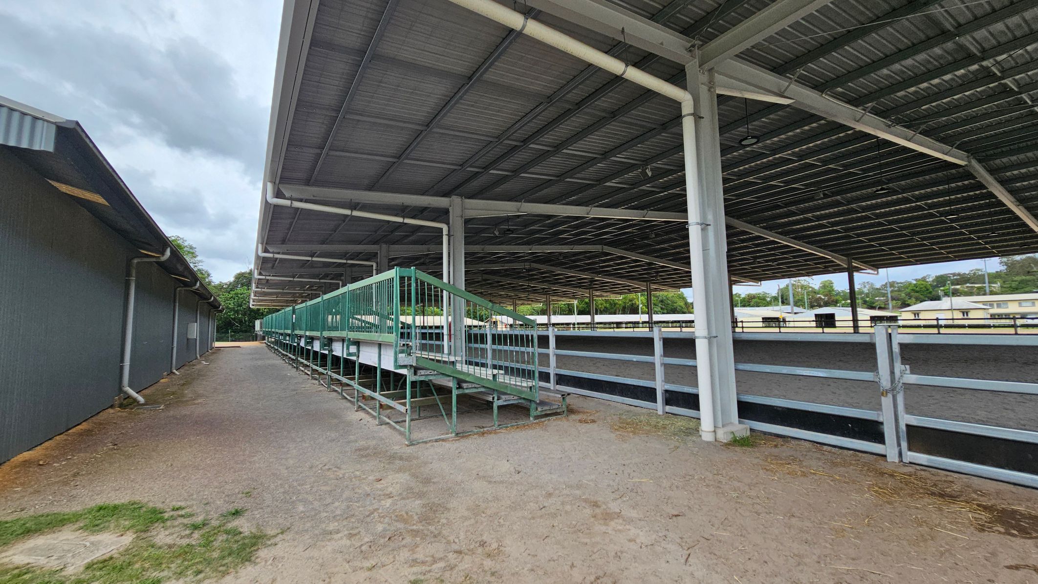 A covered outdoor area with a large metal roof supported by white pillars. On the right, there is a fenced area with a green metal ramp and seating. The ground is a mix of dirt and gravel. On the left, a long, dark building with a sloped roof runs parallel to the covered area. In the background, there are trees and some low buildings. A covered outdoor area with a large metal roof supported by white pillars. On the right, there is a fenced area with a green metal ramp and seating. The ground is a mix of dirt and gravel. On the left, a long, dark building with a sloped roof runs parallel to the covered area. In the background, there are trees and some low buildings.