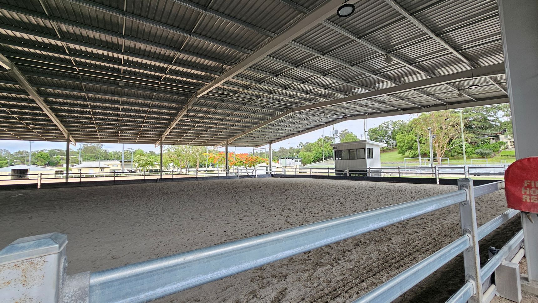 A covered equestrian arena with a metal roof and open sides. The ground is sandy, and a metal railing surrounds the area. In the background, there are trees and a small building. A red fire hose reel is visible on the right side. The ceiling has several lights and fans. A covered equestrian arena with a metal roof and open sides. The ground is sandy, and a metal railing surrounds the area. In the background, there are trees and a small building. A red fire hose reel is visible on the right side. The ceiling has several lights and fans.