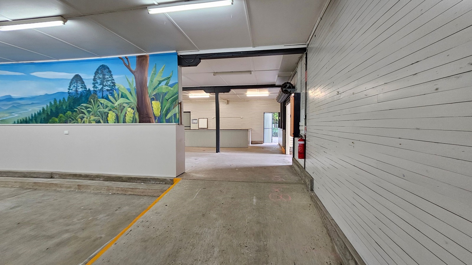 A room with a concrete floor and a white panelled ceiling. On the left, there's a mural depicting a landscape with trees and plants. In the centre, an open doorway leads to another room with a counter and a door to the outside. On the right, a white panelled wall with a fire extinguisher mounted near the doorway. Fluorescent lights are fixed to the ceiling. A room with a concrete floor and a white panelled ceiling. On the left, there's a mural depicting a landscape with trees and plants. In the centre, an open doorway leads to another room with a counter and a door to the outside. On the right, a white panelled wall with a fire extinguisher mounted near the doorway. Fluorescent lights are fixed to the ceiling.
