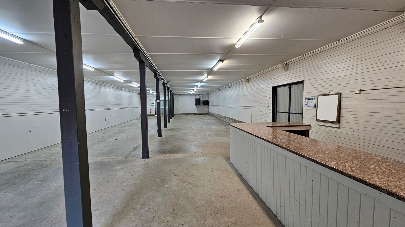A long, empty indoor space with a concrete floor and a white panelled ceiling. The left and right walls are lined with white panels. A series of black metal support beams run down the centre. At the far end, there is a double door with a metal frame and a fire extinguisher nearby. On the right, there is a small partitioned area with a notice board and a door. The lighting is provided by fluorescent tubes on the ceiling. A long, empty indoor space with a concrete floor and a white panelled ceiling. The left and right walls are lined with white panels. A series of black metal support beams run down the centre. At the far end, there is a double door with a metal frame and a fire extinguisher nearby. On the right, there is a small partitioned area with a notice board and a door. The lighting is provided by fluorescent tubes on the ceiling.