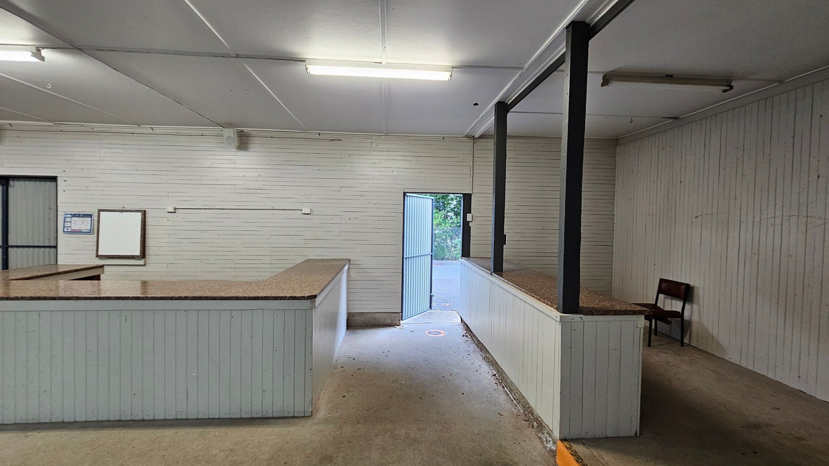A room with light green wooden walls and a white ceiling. On the left, there is a counter with a speckled brown surface. A notice board and a closed door are visible on the left wall. In the centre, an open door leads outside, showing greenery. On the right, a raised platform with a similar counter and a single red chair. The floor is concrete, and fluorescent lights are mounted on the ceiling. A room with light green wooden walls and a white ceiling. On the left, there is a counter with a speckled brown surface. A notice board and a closed door are visible on the left wall. In the centre, an open door leads outside, showing greenery. On the right, a raised platform with a similar counter and a single red chair. The floor is concrete, and fluorescent lights are mounted on the ceiling.