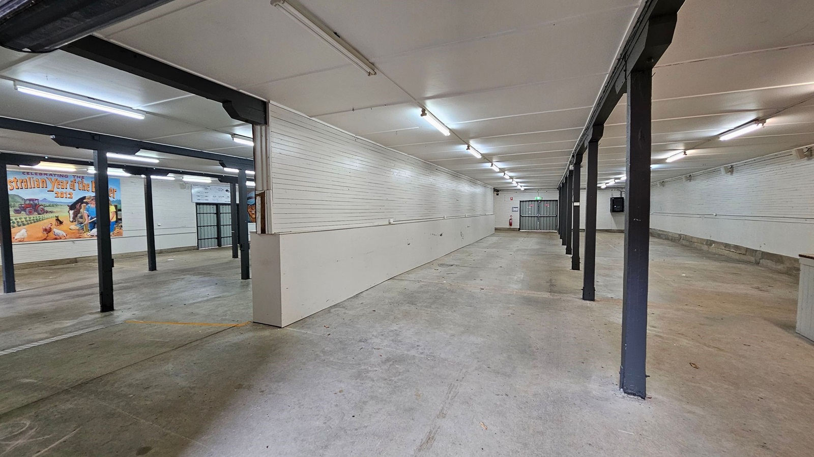 A spacious, empty indoor area with a concrete floor and a white panelled ceiling. On the left, there are black metal support beams and a mural with agricultural imagery and text. The right side features a long white wall. At the far end, a metal gate and a fire extinguisher are visible. The lighting consists of fluorescent tubes evenly spaced across the ceiling. A spacious, empty indoor area with a concrete floor and a white panelled ceiling. On the left, there are black metal support beams and a mural with agricultural imagery and text. The right side features a long white wall. At the far end, a metal gate and a fire extinguisher are visible. The lighting consists of fluorescent tubes evenly spaced across the ceiling.