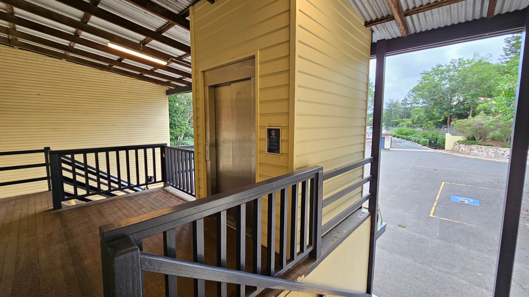A wooden ramp with black railings leads to a lift with a metallic door on the left. The structure is painted yellow with horizontal panels. The ceiling is corrugated metal with exposed beams and a fluorescent light. On the right, there is an open area with a view of a paved road and a parking space marked with a wheelchair symbol. In the background, there are green trees and a stone wall. A wooden ramp with black railings leads to a lift with a metallic door on the left. The structure is painted yellow with horizontal panels. The ceiling is corrugated metal with exposed beams and a fluorescent light. On the right, there is an open area with a view of a paved road and a parking space marked with a wheelchair symbol. In the background, there are green trees and a stone wall.