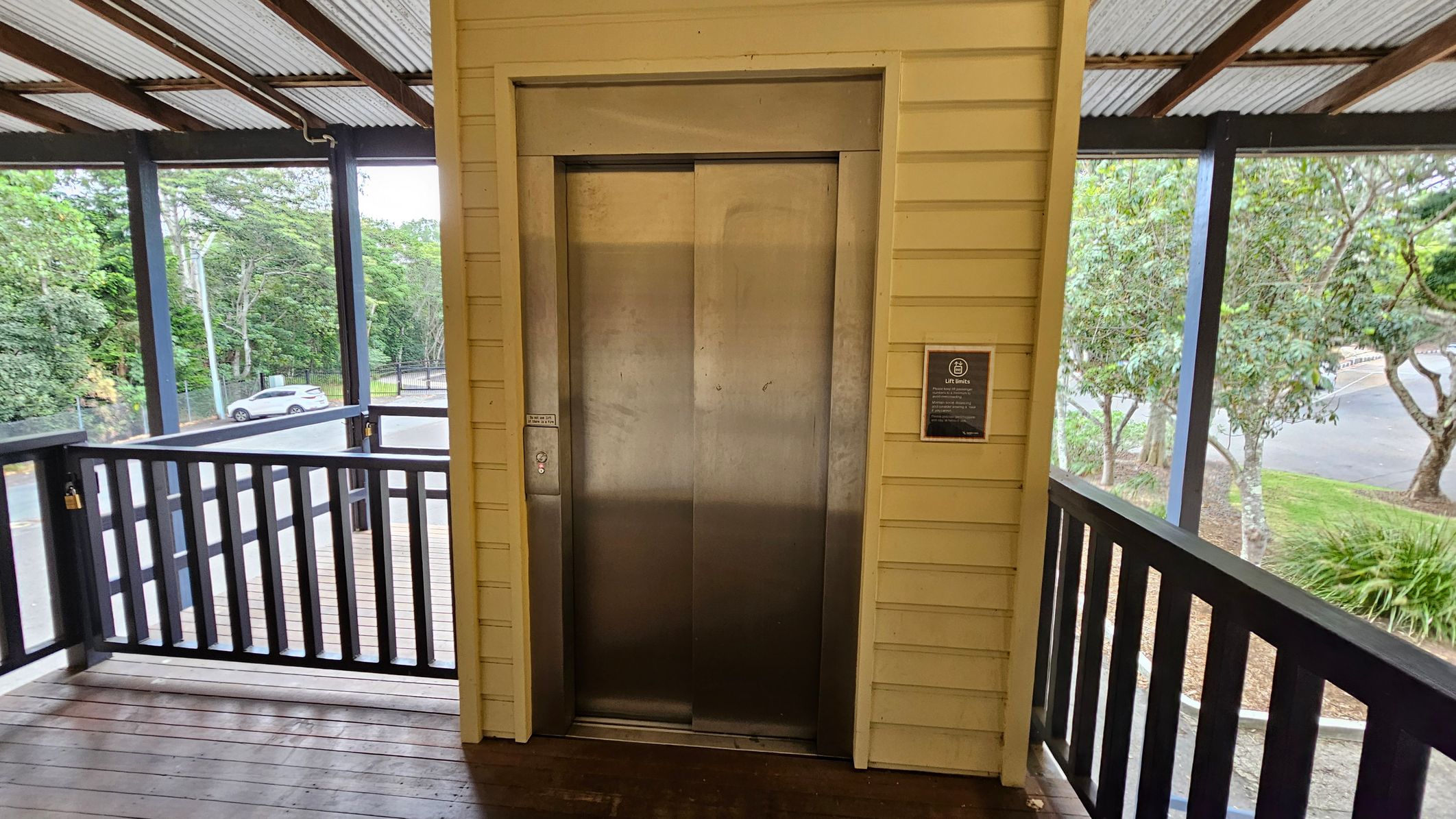 A metal lift door is centred on a wooden wall with a yellowish hue. To the right, a sign is mounted on the wall. The area is enclosed by a black railing, and beyond it, trees and a parked white car are visible. The floor is wooden, and the ceiling is made of corrugated metal with exposed beams. A metal lift door is centred on a wooden wall with a yellowish hue. To the right, a sign is mounted on the wall. The area is enclosed by a black railing, and beyond it, trees and a parked white car are visible. The floor is wooden, and the ceiling is made of corrugated metal with exposed beams.