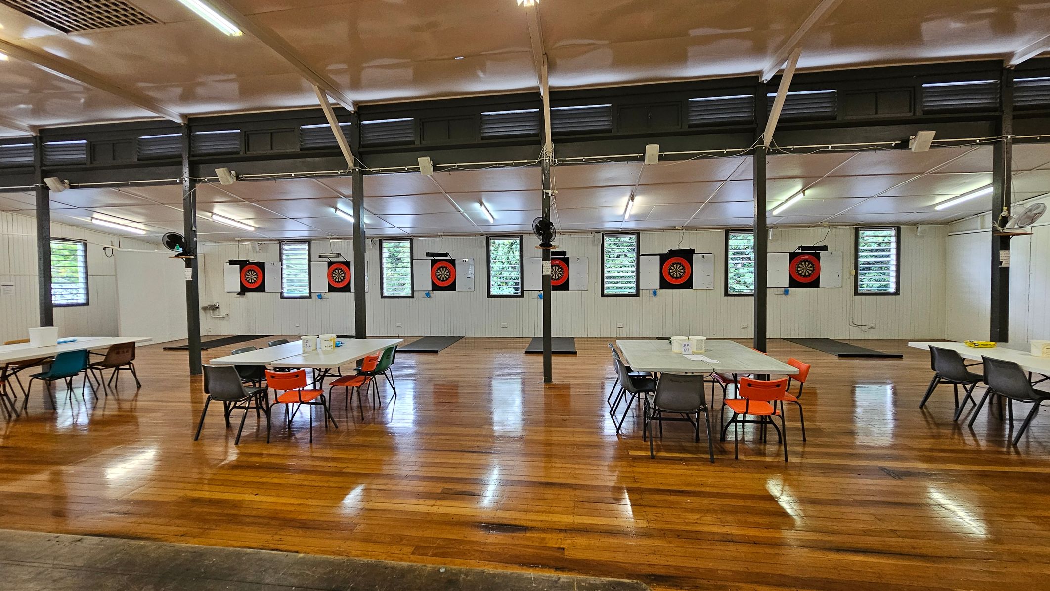 A spacious hall with polished wooden floors and a high ceiling. On the left, there are dartboards mounted on the wall, with colourful pennants hanging above. In the centre, rows of tables and chairs are arranged neatly, featuring red and grey seating. The right side has windows with louvres, allowing natural light in. A wooden plaque is mounted on the far wall. The ceiling has fluorescent lights and ceiling fans. A spacious hall with polished wooden floors and a high ceiling. On the left, there are dartboards mounted on the wall, with colourful pennants hanging above. In the centre, rows of tables and chairs are arranged neatly, featuring red and grey seating. The right side has windows with louvres, allowing natural light in. A wooden plaque is mounted on the far wall. The ceiling has fluorescent lights and ceiling fans.